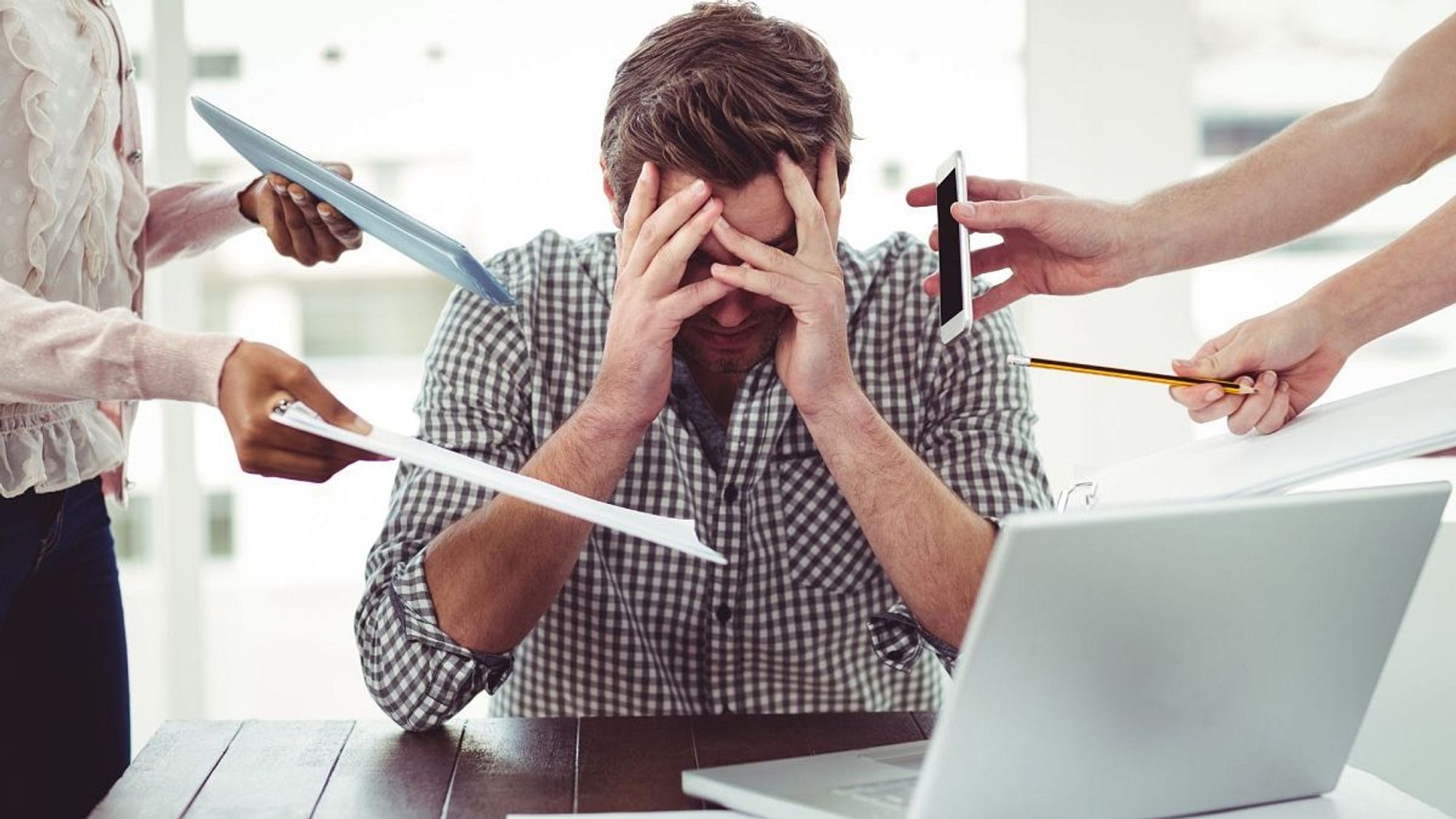 A man with his hands over his face at the desk with a computer in front of him surrounded by hands who seem to give him work to do