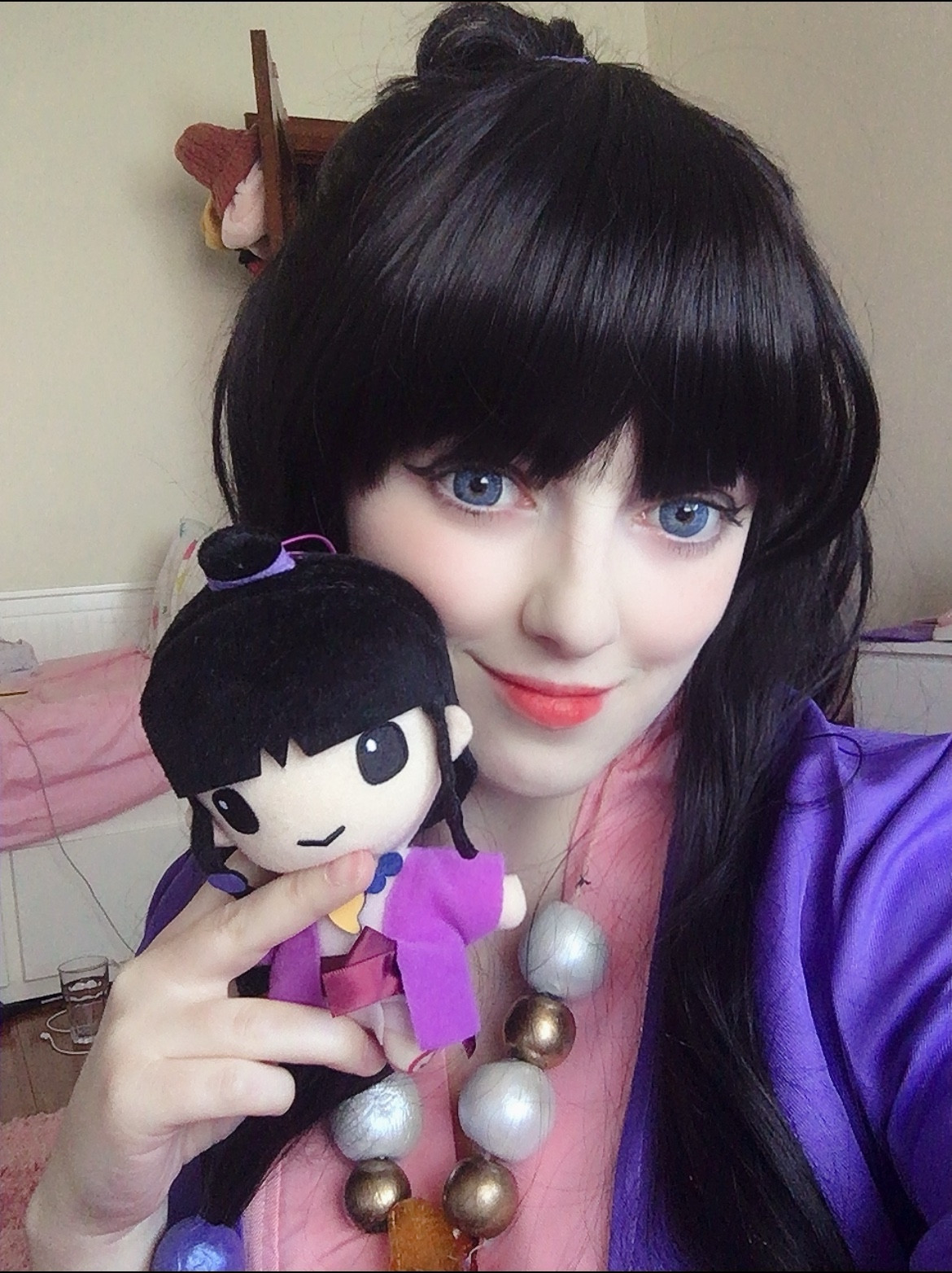 cosplay of maya fey from ace attorney. they have long black hair, a purple robe and a magatama necklace. they are holding a plush toy of maya as well like the world's most awesome twins 