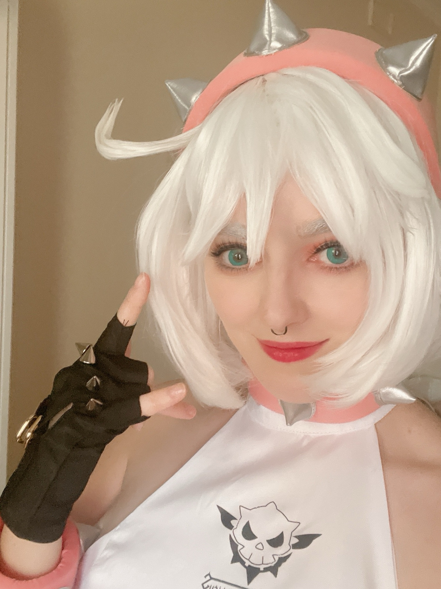 cosplay of elphelt valentine from guilty gear. they have short white hair and giving the "rock on" symbol with their hand. they also have a spiked pink headband and choker, but not the jacket because it was too hot outside