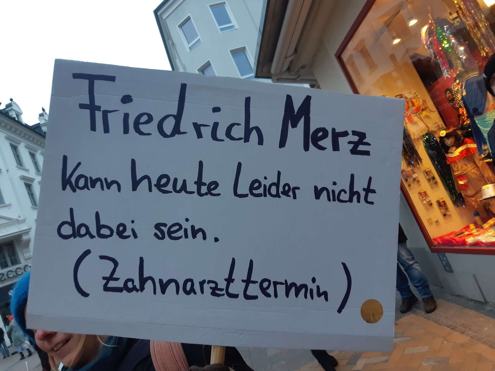 Demo-Schild mit Aufschrift "Friedrich Merz kann heute leider nicht dabei sein. (Zahnarzttermin)"