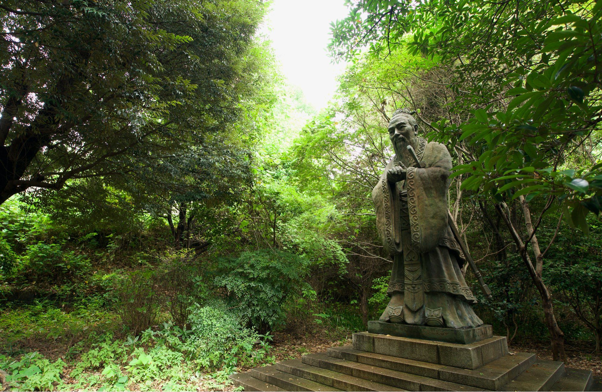 A statue of Confucius, standing on a stepped pedestal, is on the right third of the frame. The framing of the statue is him looking down and towards the left. Green flora fills the majority of the background