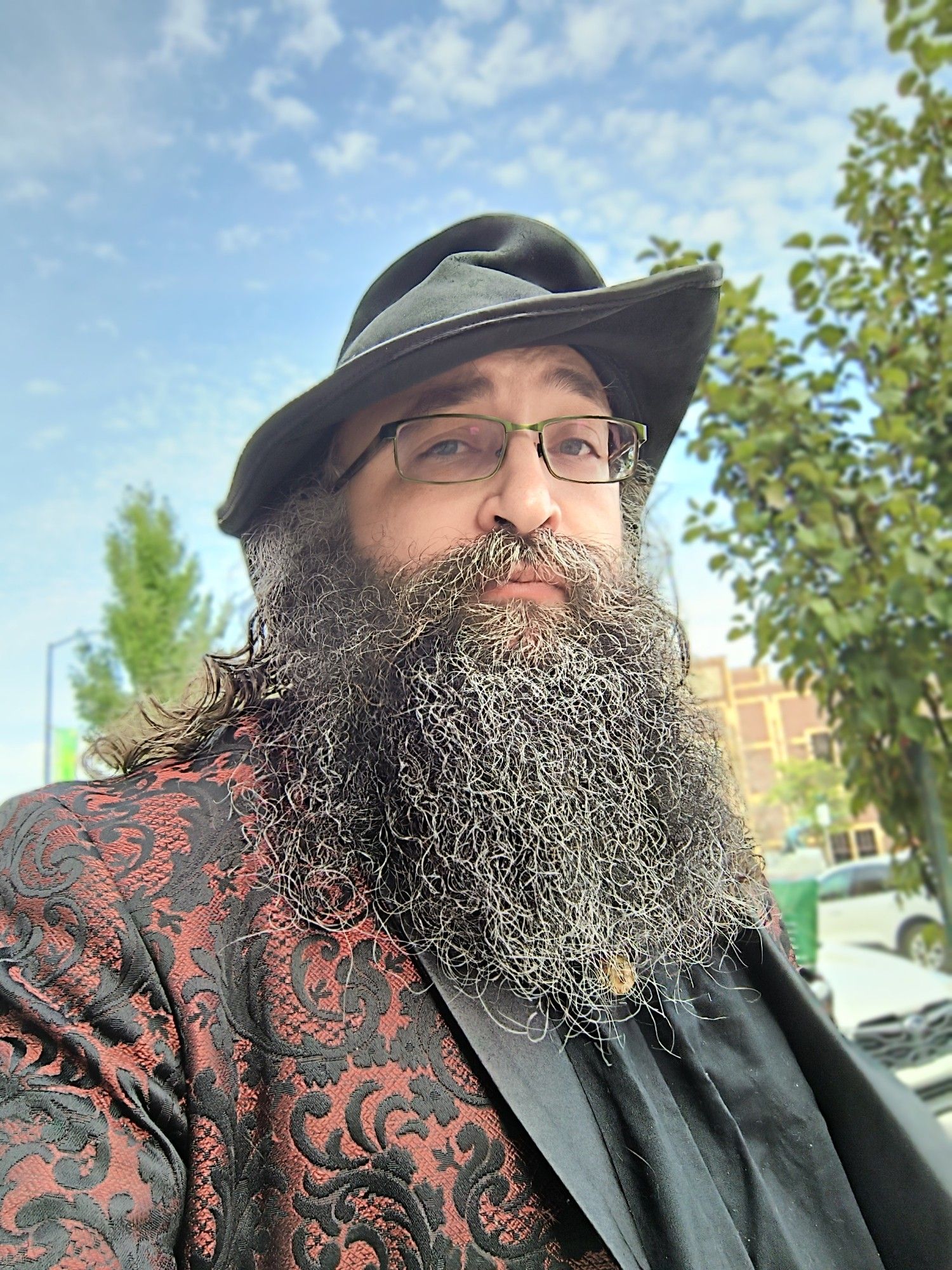 Bearded caucasian male-presenting individual below a blue sky. Fancy suitcoat and a green hat being worn, as well as a black shirt.