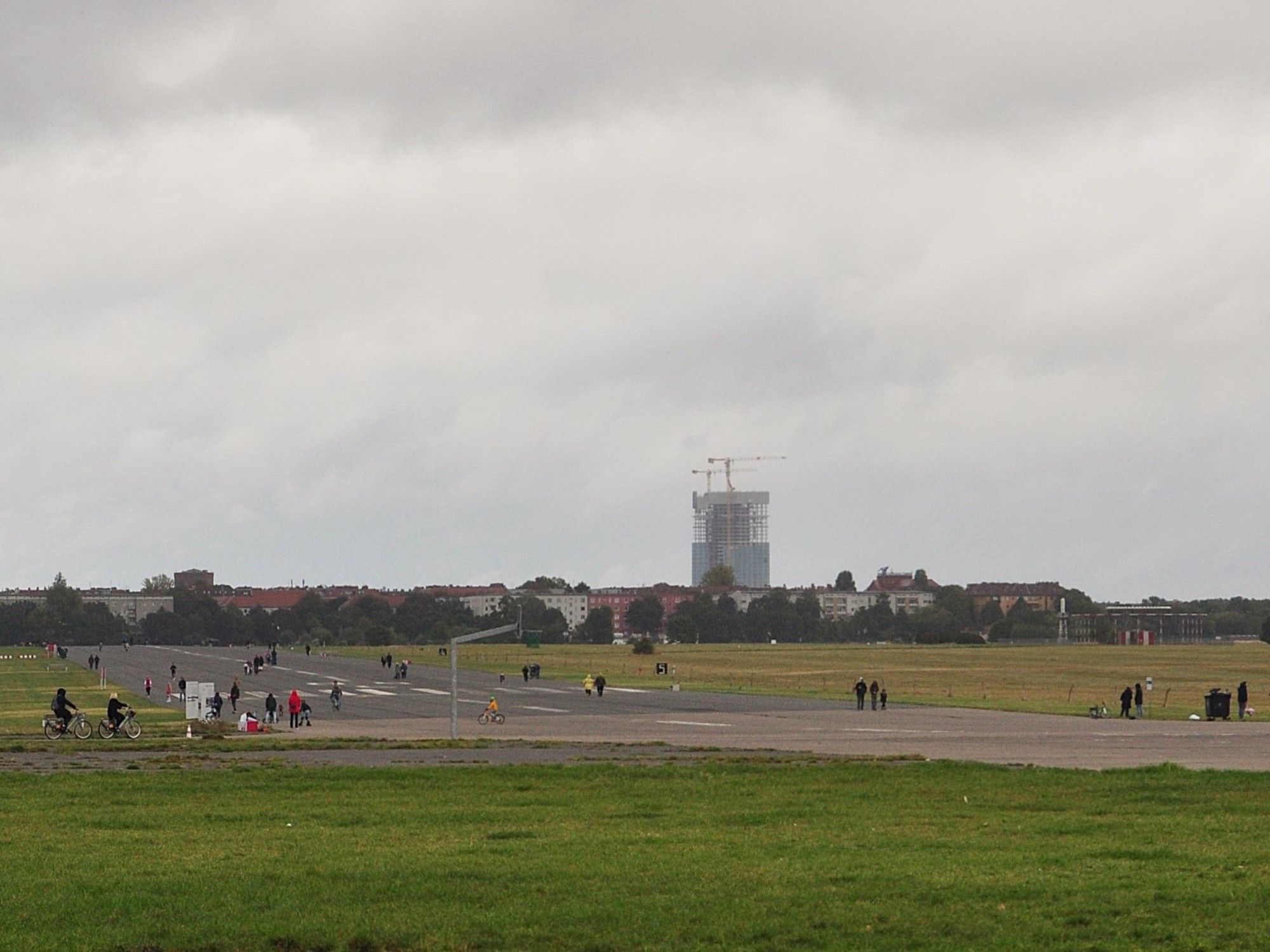 tempelhofer feld, eine große freifläche, wiese mit einer start- und landebahn. überall paar wenige leute. hinten am anderen ende des feldes häuser mit bäumen und ein hochhaus im bau mit kran obendrauf.