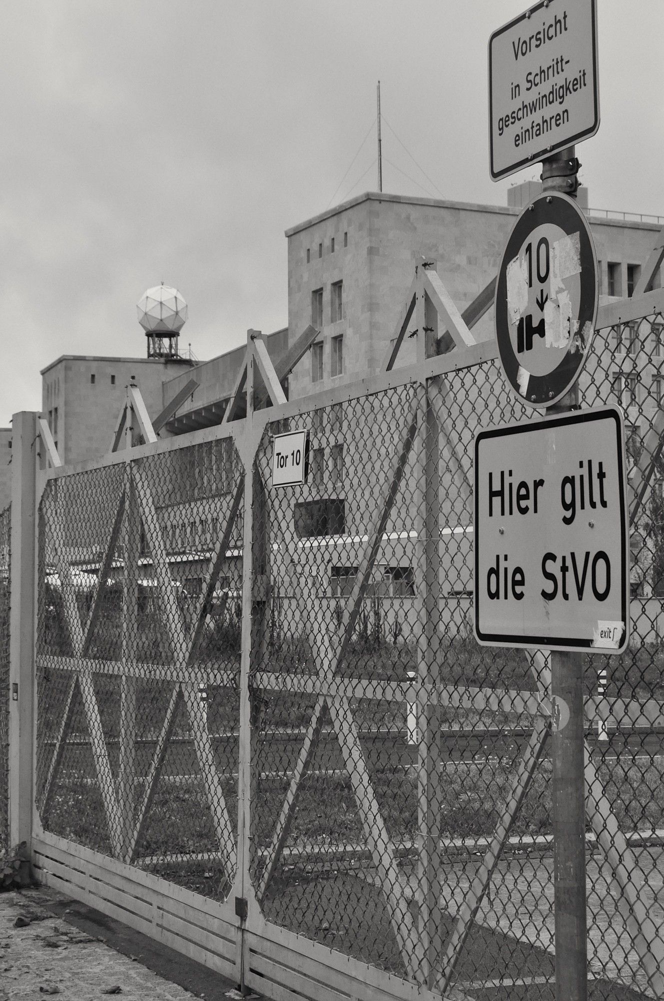 s/w. metallenes zauntor am gebäude des nazistischen tempelhofer flughafens. so eine radar-kugel auf dach. straßenschilder "hier gilt die stvo" und andere.