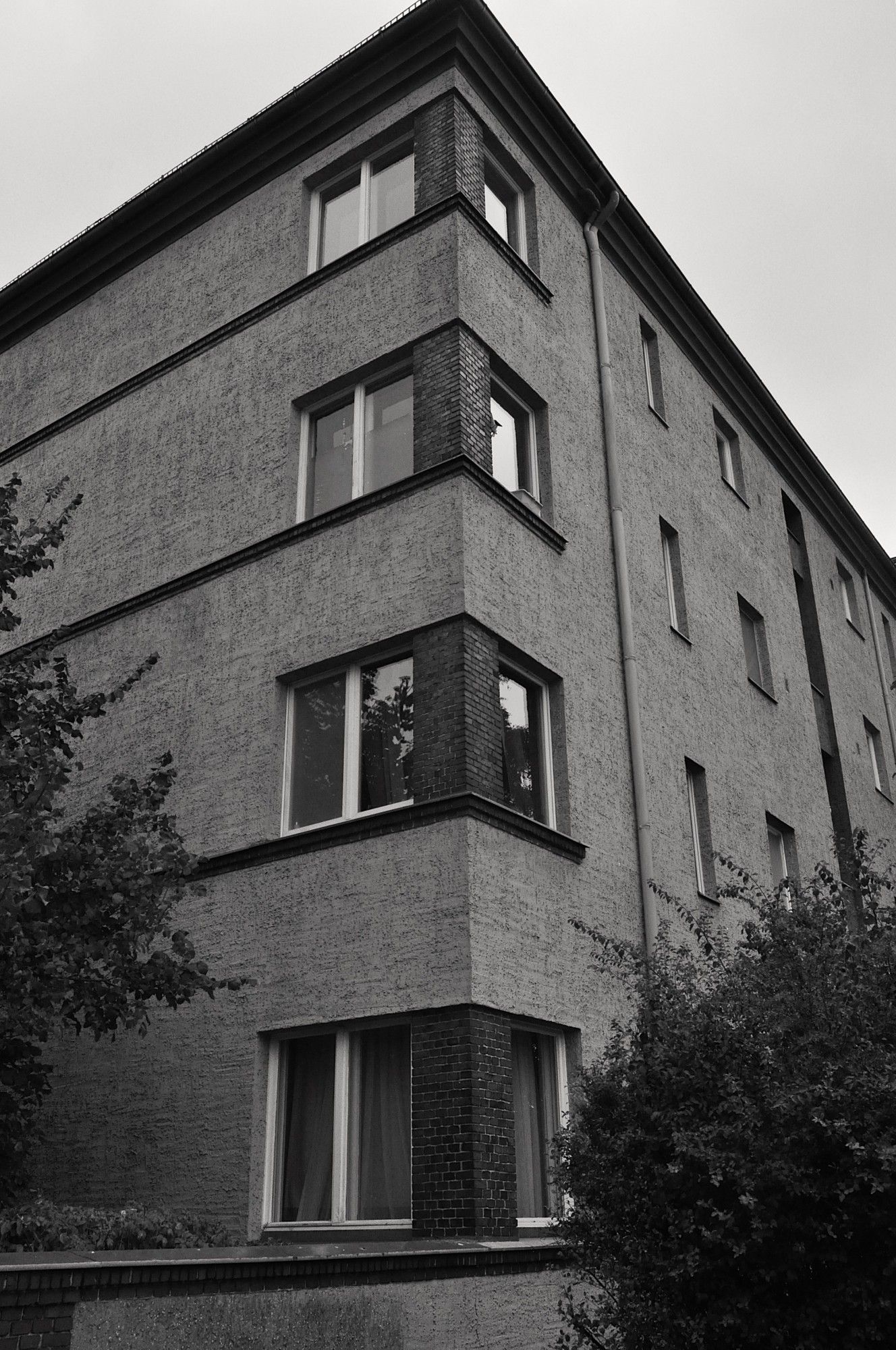 s/w. dreistöckiges haus auf die ecke fotografiert. teilweise verputzte wand, aber an der eck-kante backsteine zu sehen und fenster. grauer himmel.