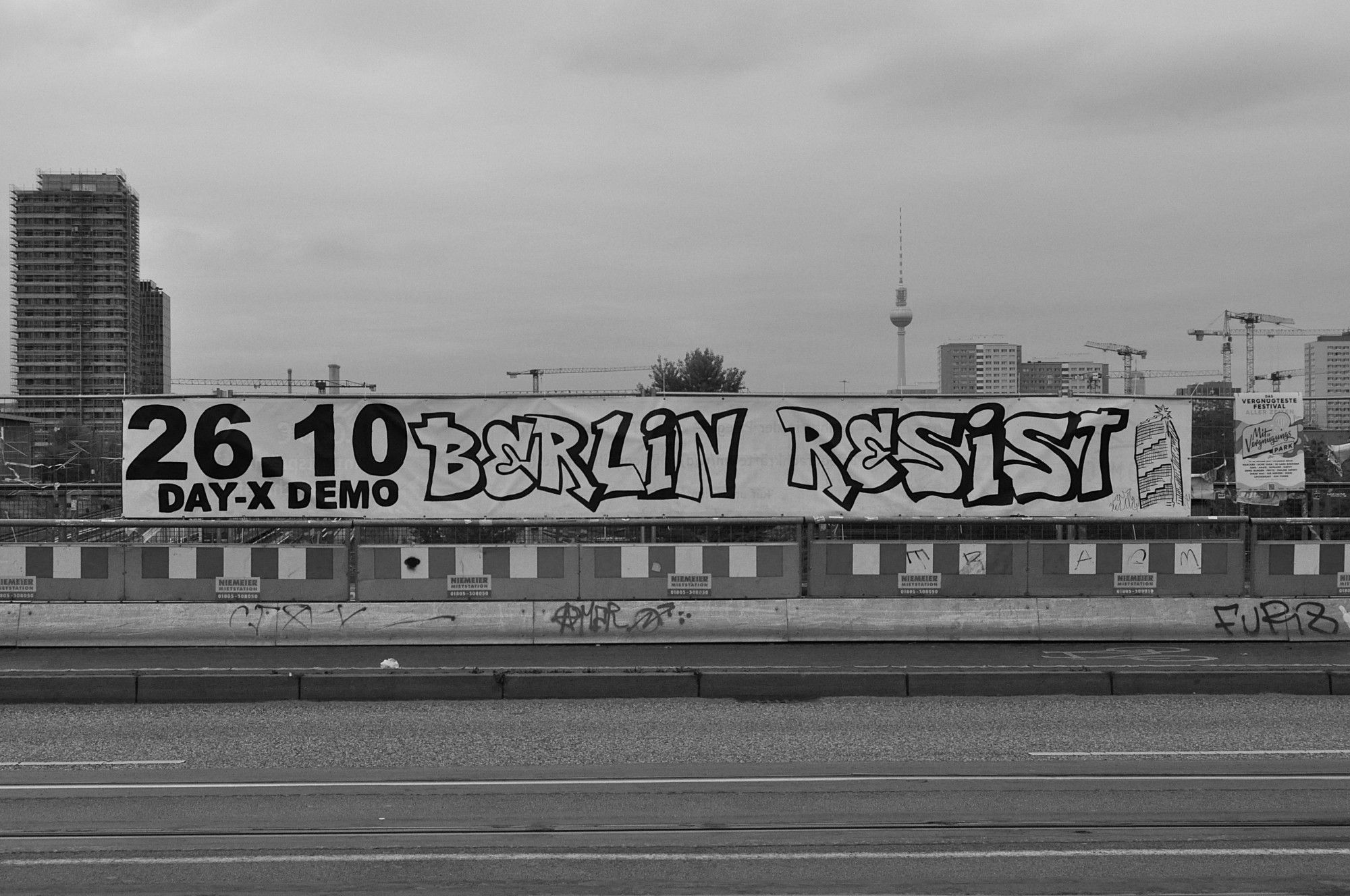 s/w. Großes banner an bauzaun auf der warschauer brücke. dahinter berlin mit hochhaus in bau, vielen kränen und dem fernsehturm. auf dem banner: "26.10. day-x-demo BERLIN RESIST".