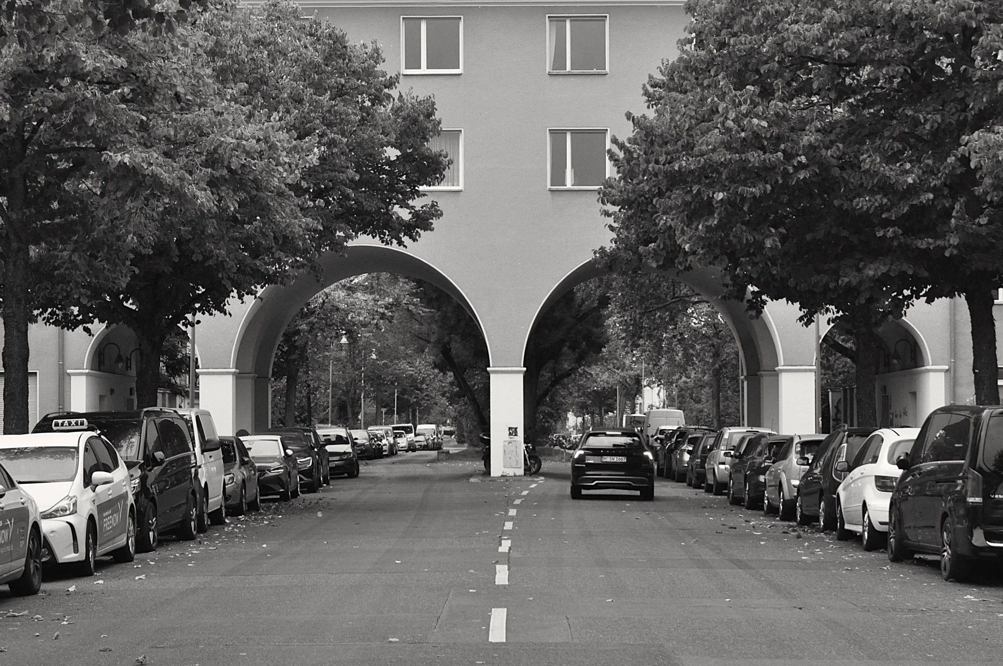 s/w. zweispurige straße geht in zwei bögen unter einem haus durch, drüber noch 2 stockwerke. für die gehwege eigene kleine tunnel. rechts und links am bildrand je ein baum. ein auto auf der rechten spur mit bremslichtern an.