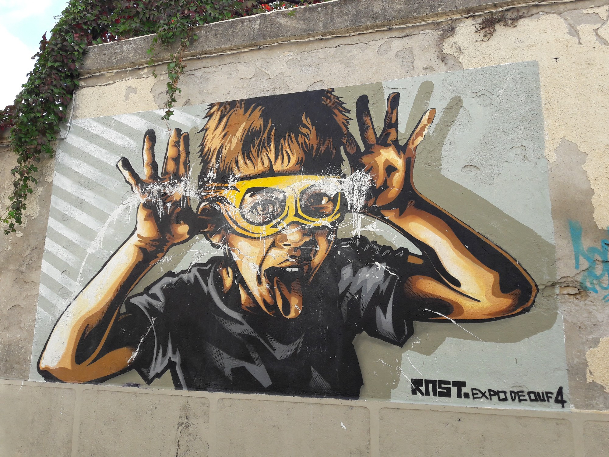 Sur un mur dans la rue, un dessin de Rust est collé : le buste d'un enfant avec des lunettes jaunes qui tire la langue les mains écartées derrière les oreilles