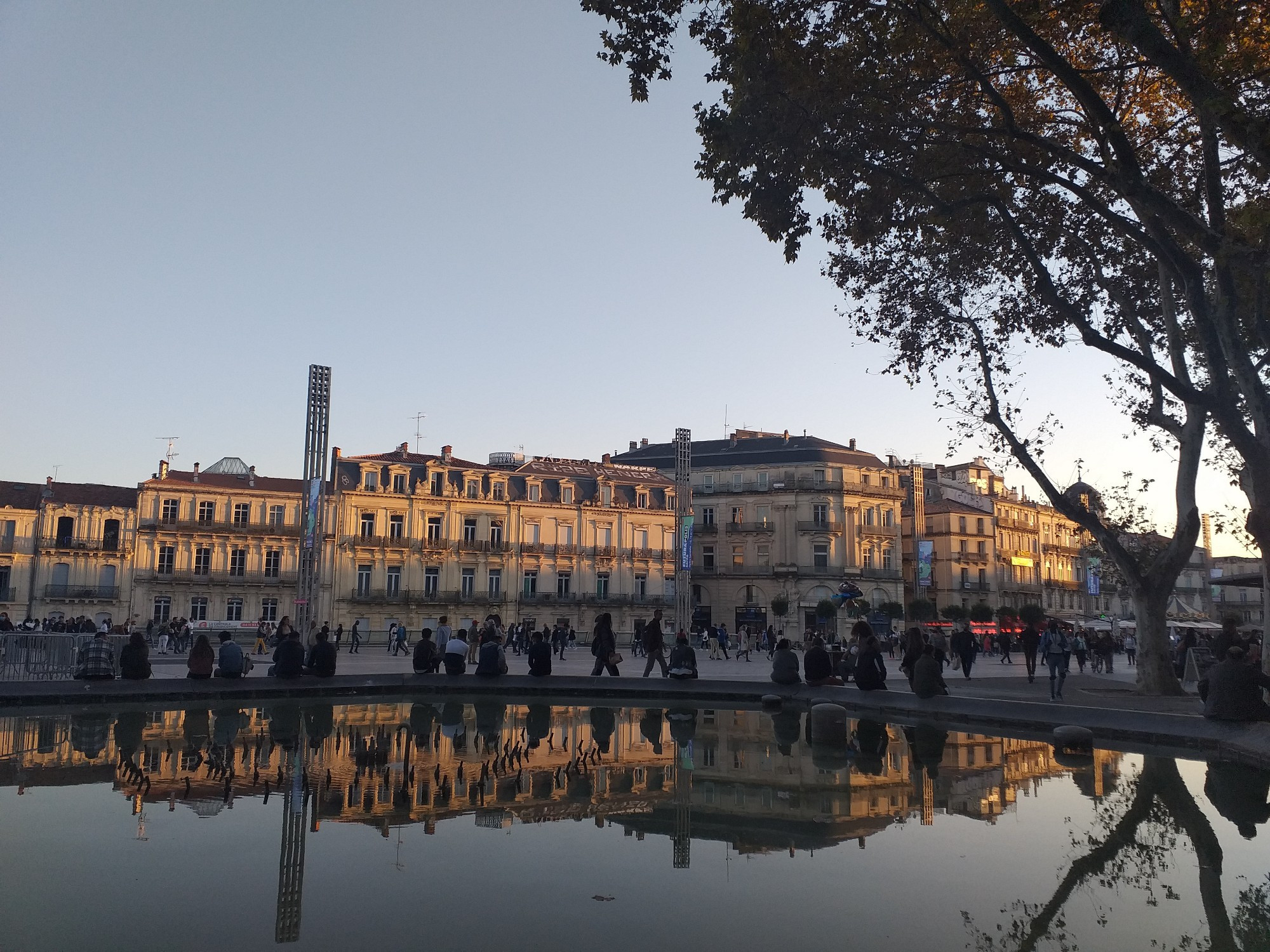 Au premier plan, une étendue d'eau dans laquelle se reflètent des branches d'arbre  des gens assis sur un muret (le bord de la fontaine aujourd'hui disparue) et les immeubles de type haussmanien de la place en arrière plan. 