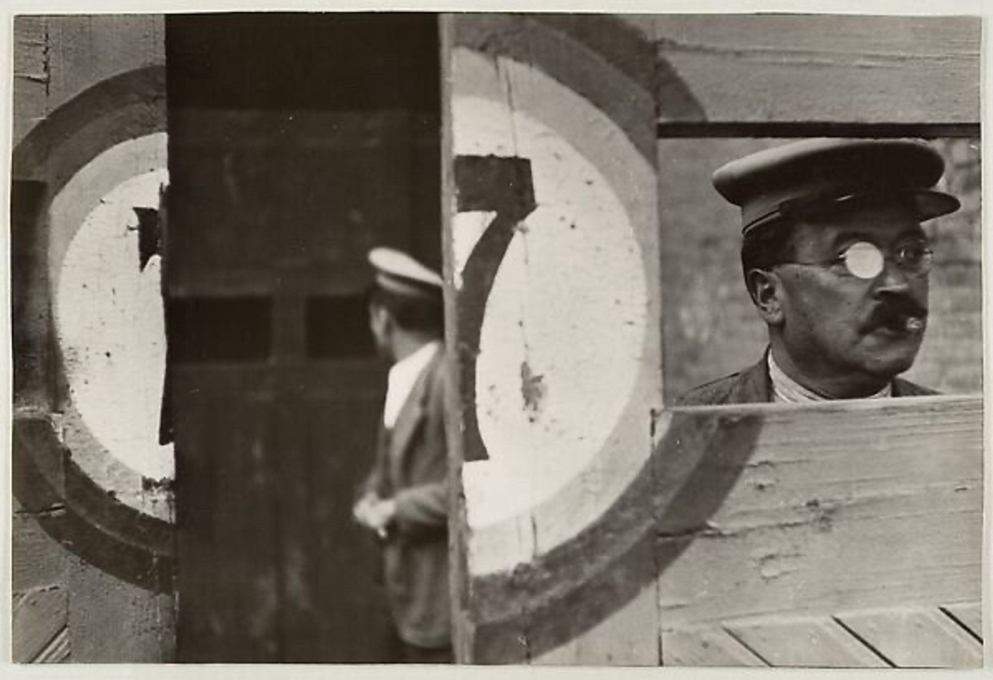 A photo by Henri Cartier Bresson from 1933 in Valencia, Spain, showing the inside of a bullring. An attendant wearing glasses and a cap looks out of a rectangular opening in a wooden gate, painted with the number 7. Another attendant stands out of focus in the background