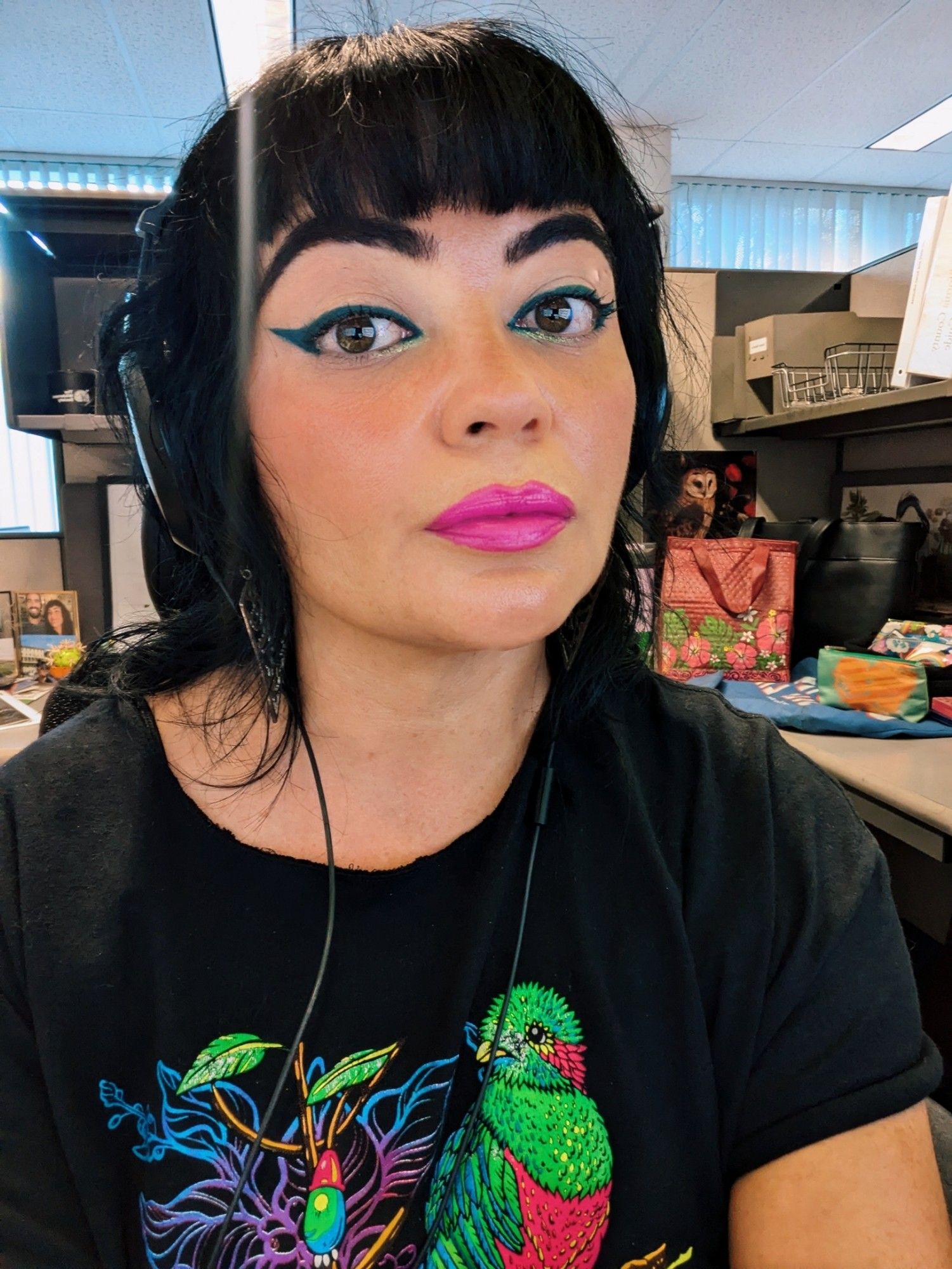 A picture of me, a female presenting person, sitting in a cubicle with a blank expression. I'm wearing large over the ear headphones. I have long black hair with straight bangs, and I'm wearing teal cateye eyeliner, with some shimmer in the inner corners, and neon pink lipstick. My shirt is an oversized black t shirt with a neon resplendent quetzal on it.