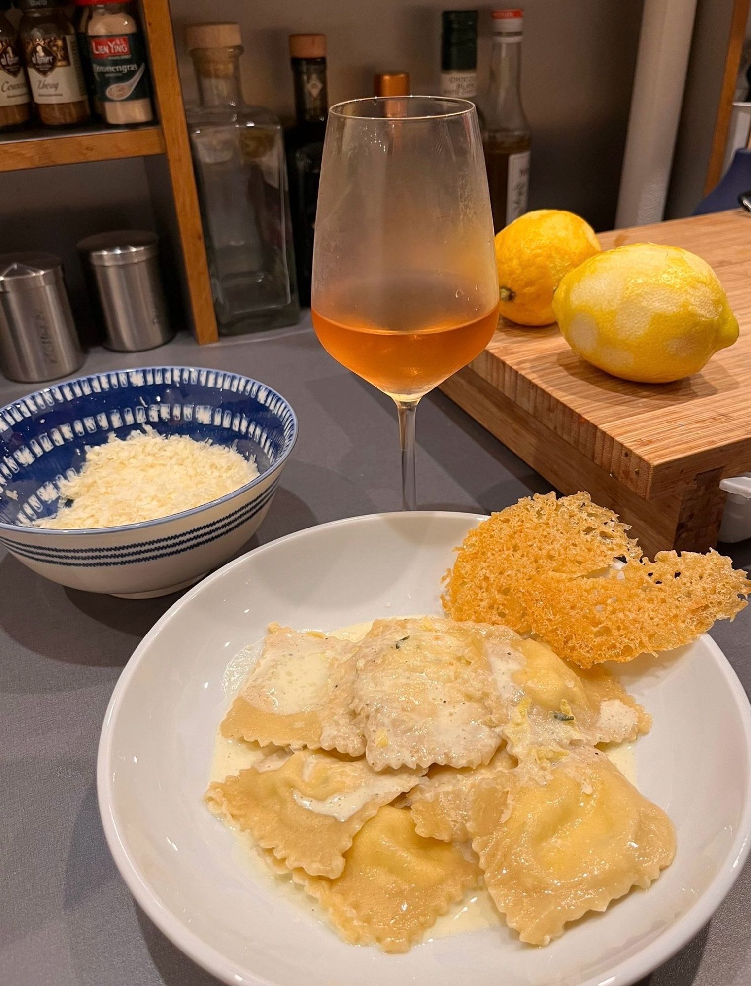 Ein weißer tiefer Teller mit Ravioli (Ricotta Zitrone Thymian) auf einem Zitronen- Thymian- Schaum und Parmesanchip. Eine blauweiße Schale mit geriebenen Parmesan, ein beschlagenes Glas Rosé, zwei Zitronen auf einem Holzbrett