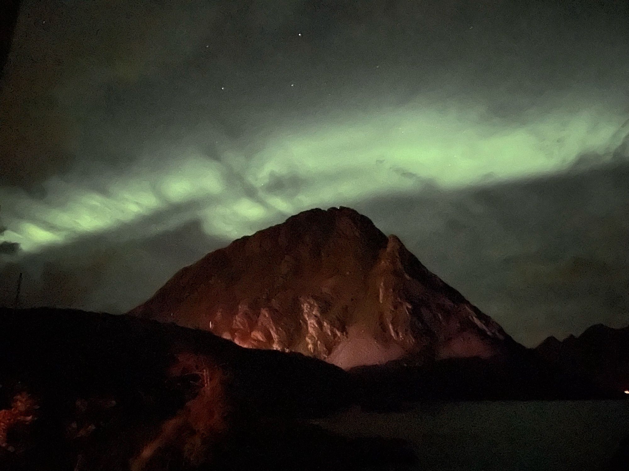 Grünlich leuchtende Streifen Nordlicht an Nachthimmel. Es scheint durch dünne Schleierwolken. Darunter die Silhouette eines Berges