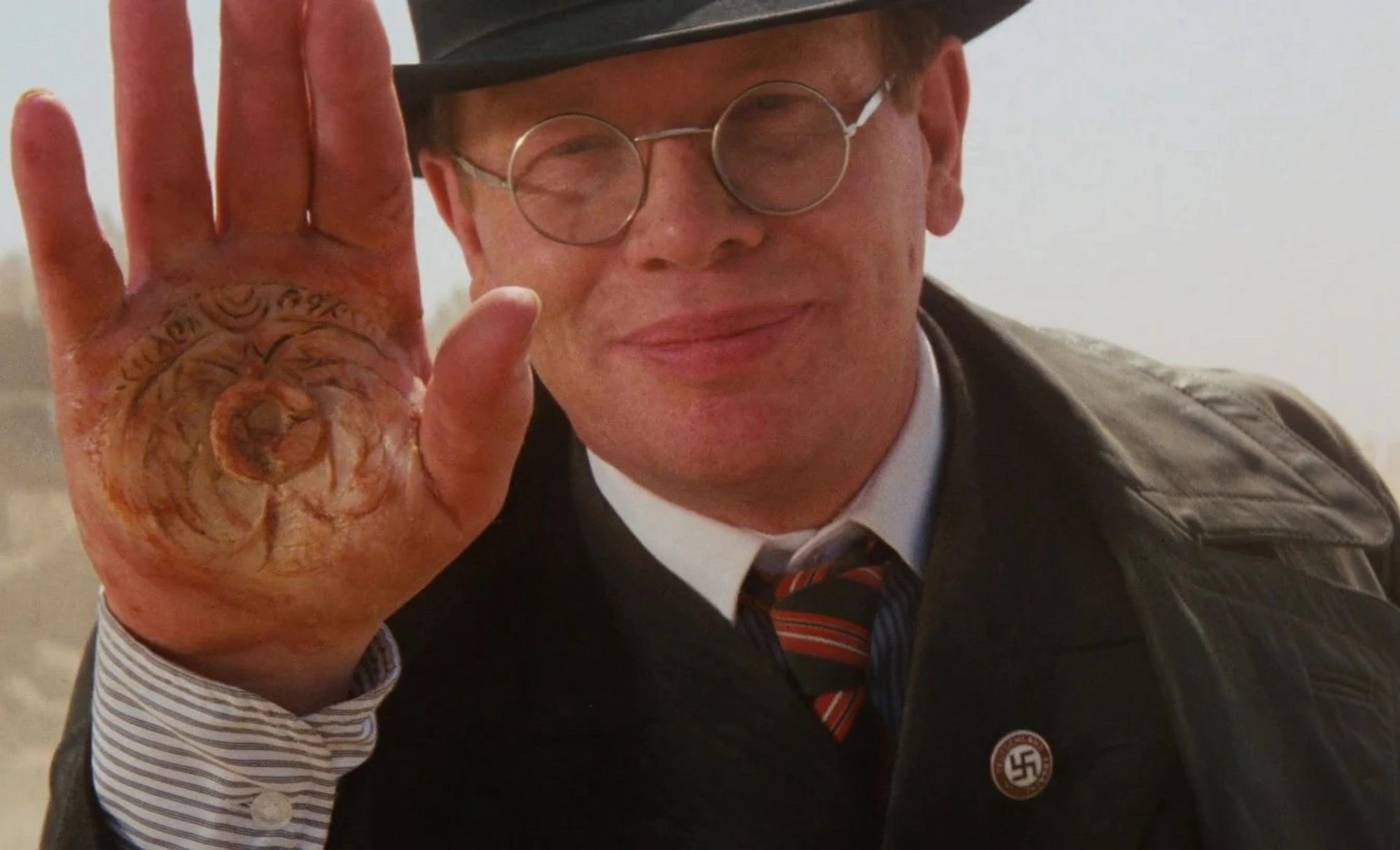 frame from the movie Raiders of the Lost Ark where the bespectacled toady Nazi lifts up his severely burns palm to reveal the imprint of a medallion that will supposedly reveal important information on where the Ark is hidden (if I have this plot point wrong, don’t tell me)