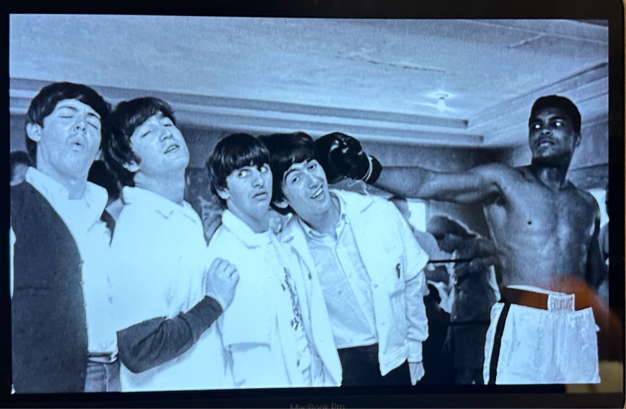 B&W photo of a young Muhammed Ali, right, (then Cassius Clay) in his practice ring where he was training to fight Sonny Liston in Miami Beach, FL, 1964, extending his right arm (viewer left) in a mock punch, and The Beatles playing along. The boxing glove touches George Harrison’s head first, knocking into Ringo Starr. Ringo breaks the chain reaction but first John Lennon then next Paul McCartney, in line with the other band members, play act along with the pretend punch