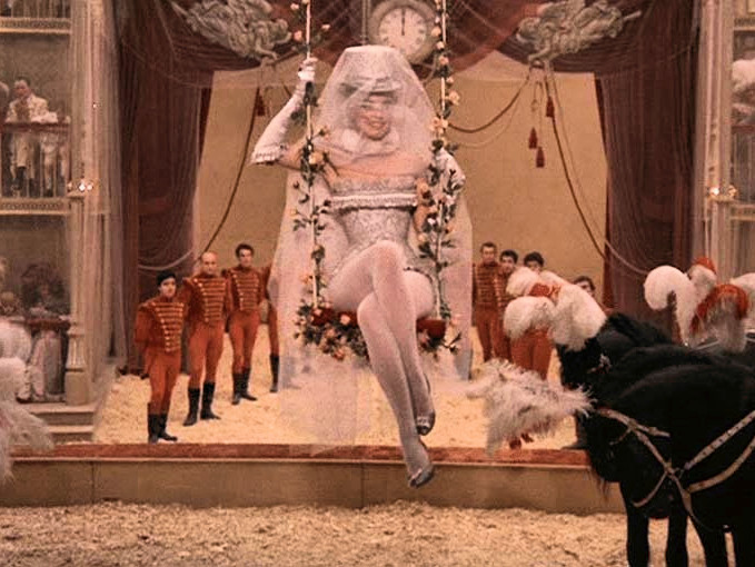 circus scene from Juliet of the Spirits featuring a woman on a trapeze wearing a white top hat and veil hanging above a group of men dressed as ringmasters and black plumed horses that are bowing to her