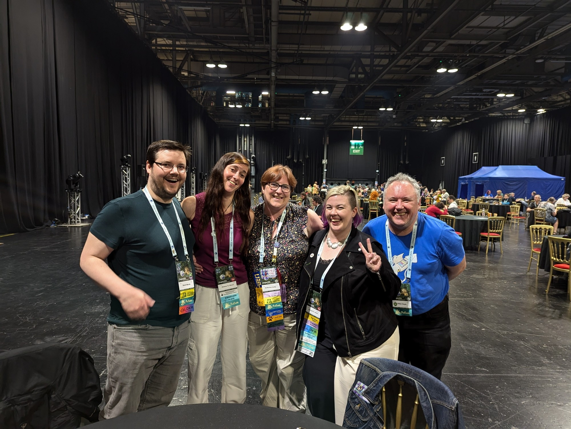 Five Escape Artist podcasters, past and present, stand together in a large convention center. Each wears a lanyard covered in colorful ribbons