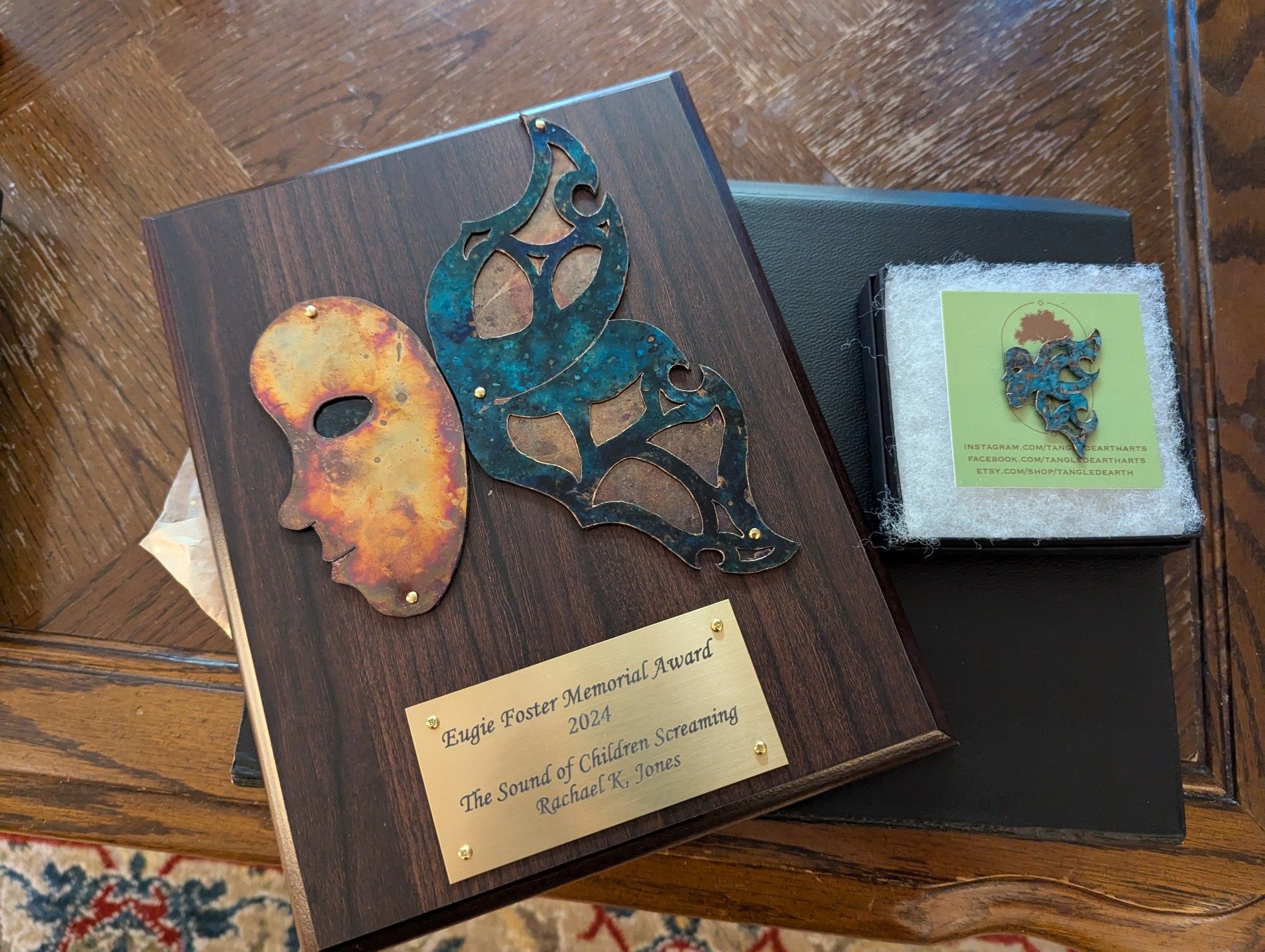 The Eugie Foster Memorial Award: A beautiful metalwork mask with wings mounted on a wooden plaque. It is engraved with the following text:
Eugie Foster Memorial Award 2024
"The Sound of Children Screaming"
Rachael K. Jones

A small metalwork pin sits beside the plaque, featuring the same mask design.