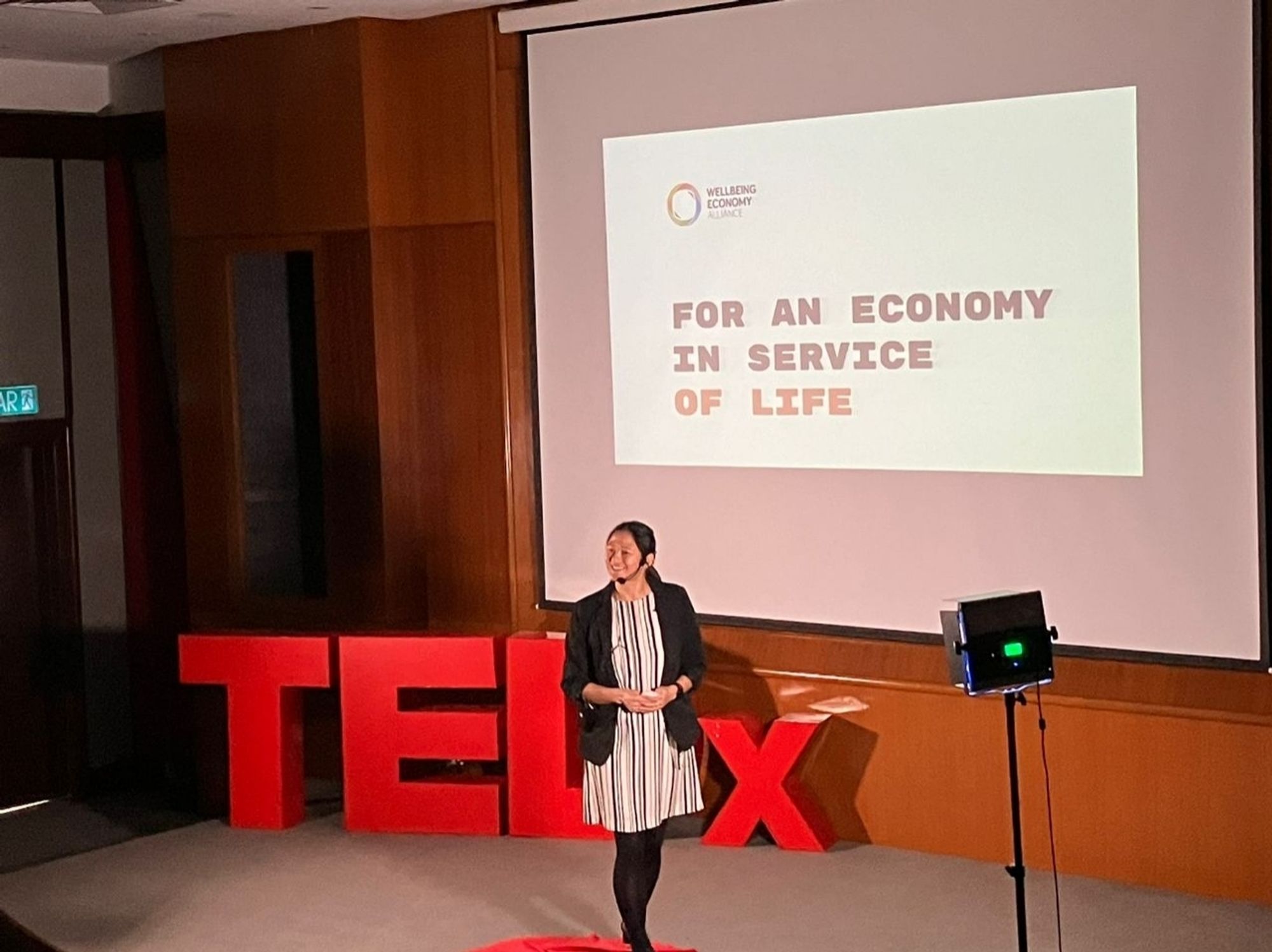 Photo of me on stage on a red circle. Behind me are large red letters which spell out TEDx. On the screen behind me it says "FOR AN ECONOMY IN SERVICE OF LIFE"