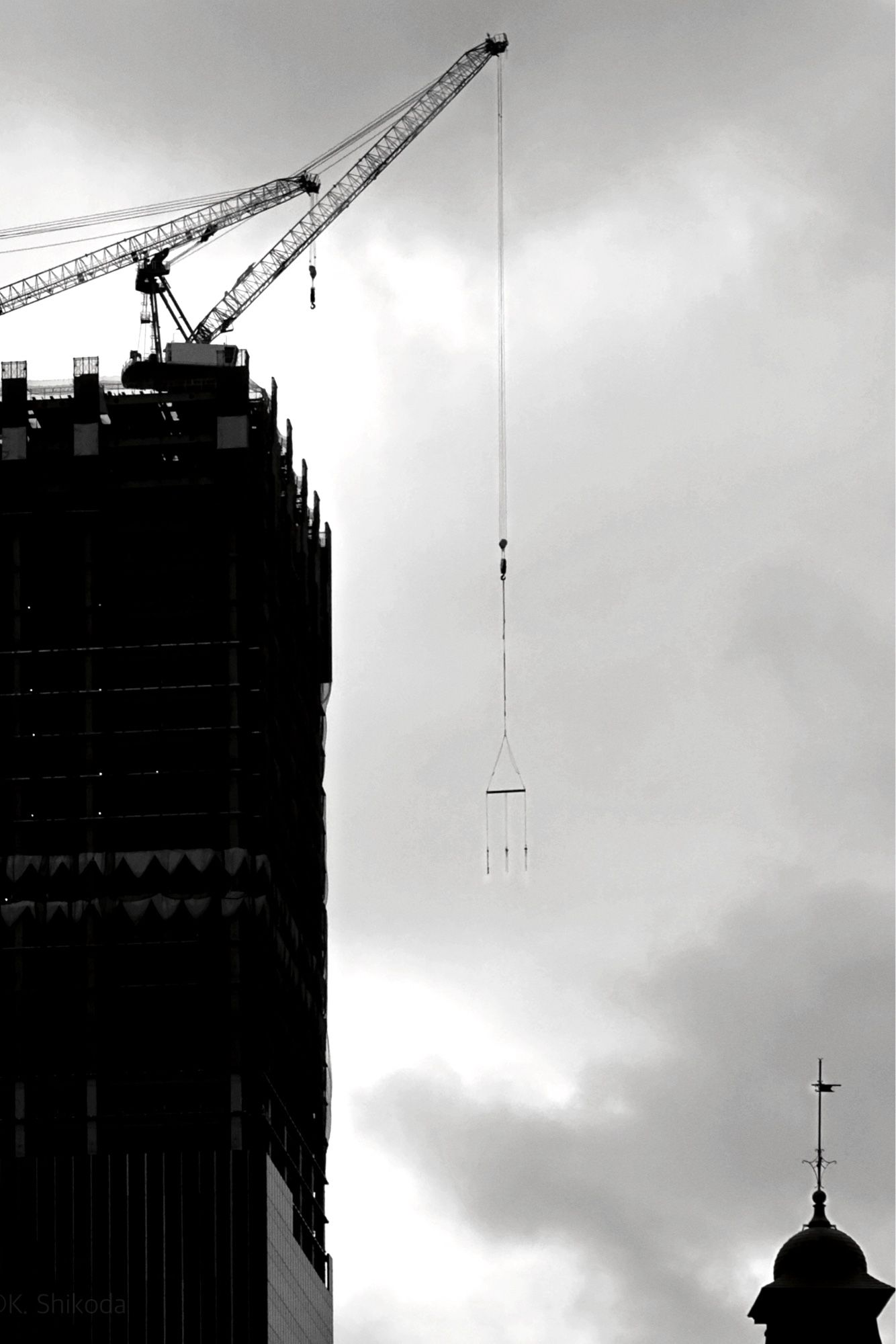 Vertical black and white photograph. Under a cloudy sky, a skyscraper under construction can be seen on the left, with two cranes visible on its roof. One of them has a horizontal bar with three wires hanging from it for hanging something. At the bottom right, one of the iconic towers attached to the roof of Tokyo Station can be seen.

縦位置のモノクロ写真
曇り空が広がる中、左に建設中の超高層ビルがあり、その屋上には2本のクレーンが見える。そのうちの一本から何かをぶら下げるための三本のワイヤーが垂れ下がった横棒が吊るされている。右下には東京駅の屋根に取り付けられた象徴的なタワーの一つが見えている。