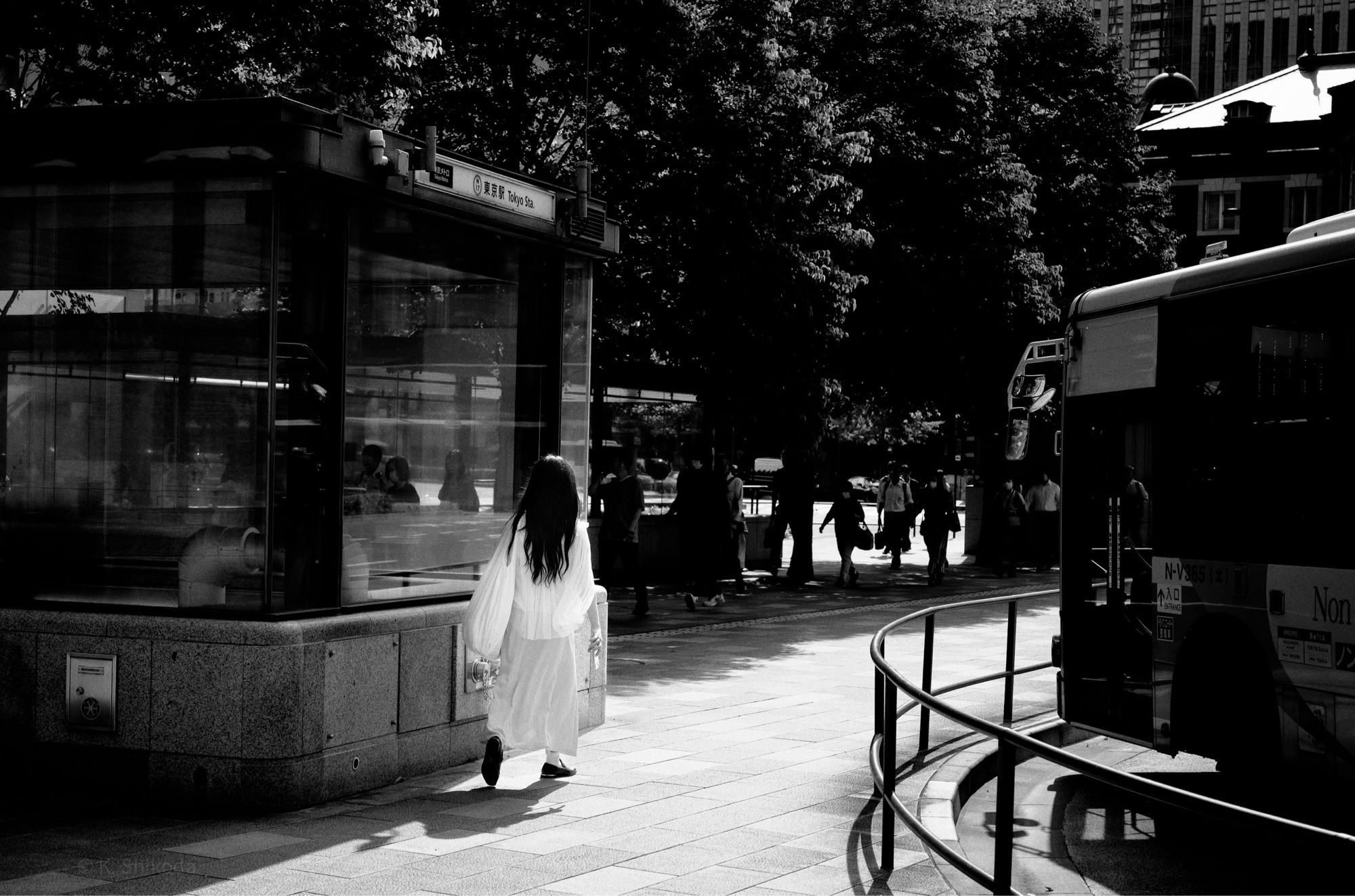 Monochrome photo
A woman in a white dress is walking in the sunlight near the Marunouchi exit of Tokyo Station, in the opposite direction to the workers, toward Tokyo Station. All the workers are walking in the shade of the trees to avoid the sun. Only she, dressed in white, is in the sunlight. A bus is parked on the right.

モノクロ写真
東京駅丸の内口の近くで、日差しを浴びながら白い服の人が、ワーカーとは逆方向の東京駅に向かって歩いている。ワーカーは皆日差しを避けて木陰を歩いている。白い服の彼女だけが日差しの中にいる。右側にはバスが停まっている。