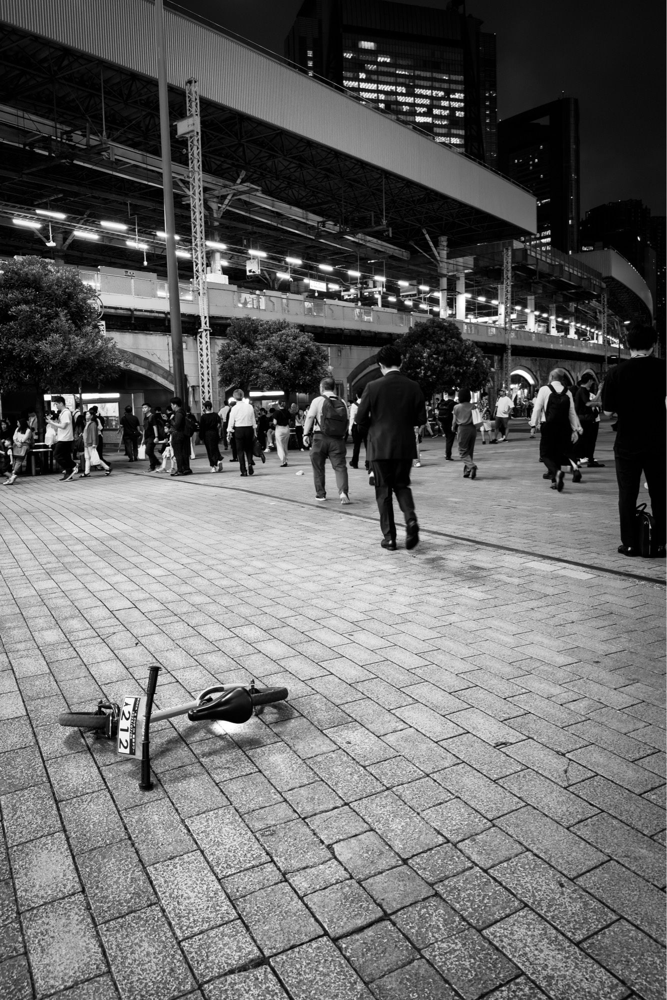 モノクロ写真。東京のサラリーマンが帰宅する時間帯の新橋駅前に、何故か子供用のバランスバイクが乗り捨てられている。

A black and white photo of a child's balance bike abandoned in front of Shinbashi Station at a time when Tokyo office workers are heading home.