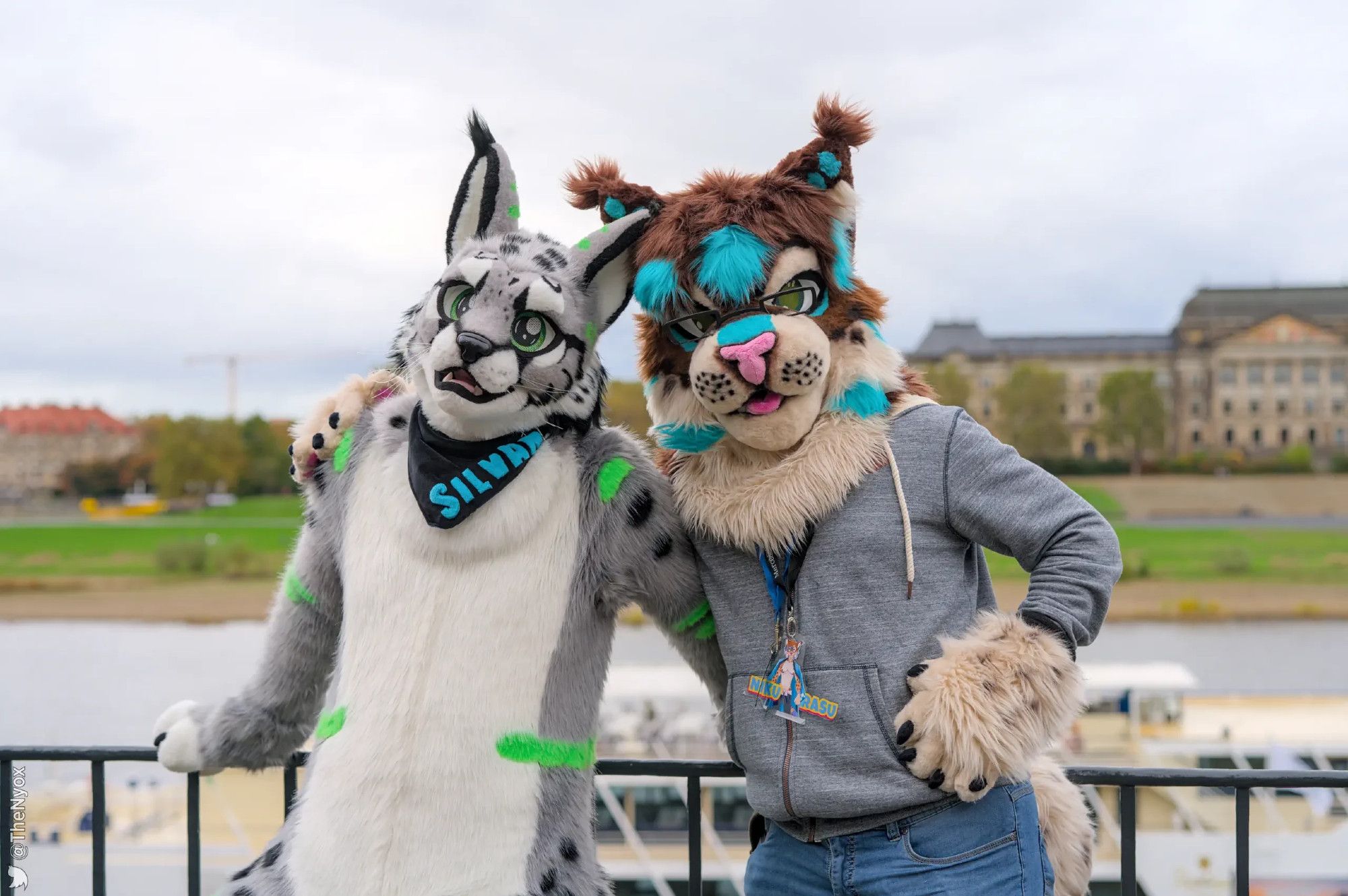 Me in my partial fursuit (a brown lynx with light blue accents) besides Silvan in his fullsuit (a silver lynx with green accents). We are standing in front of the Elbe, a river in the inner city of Dresden