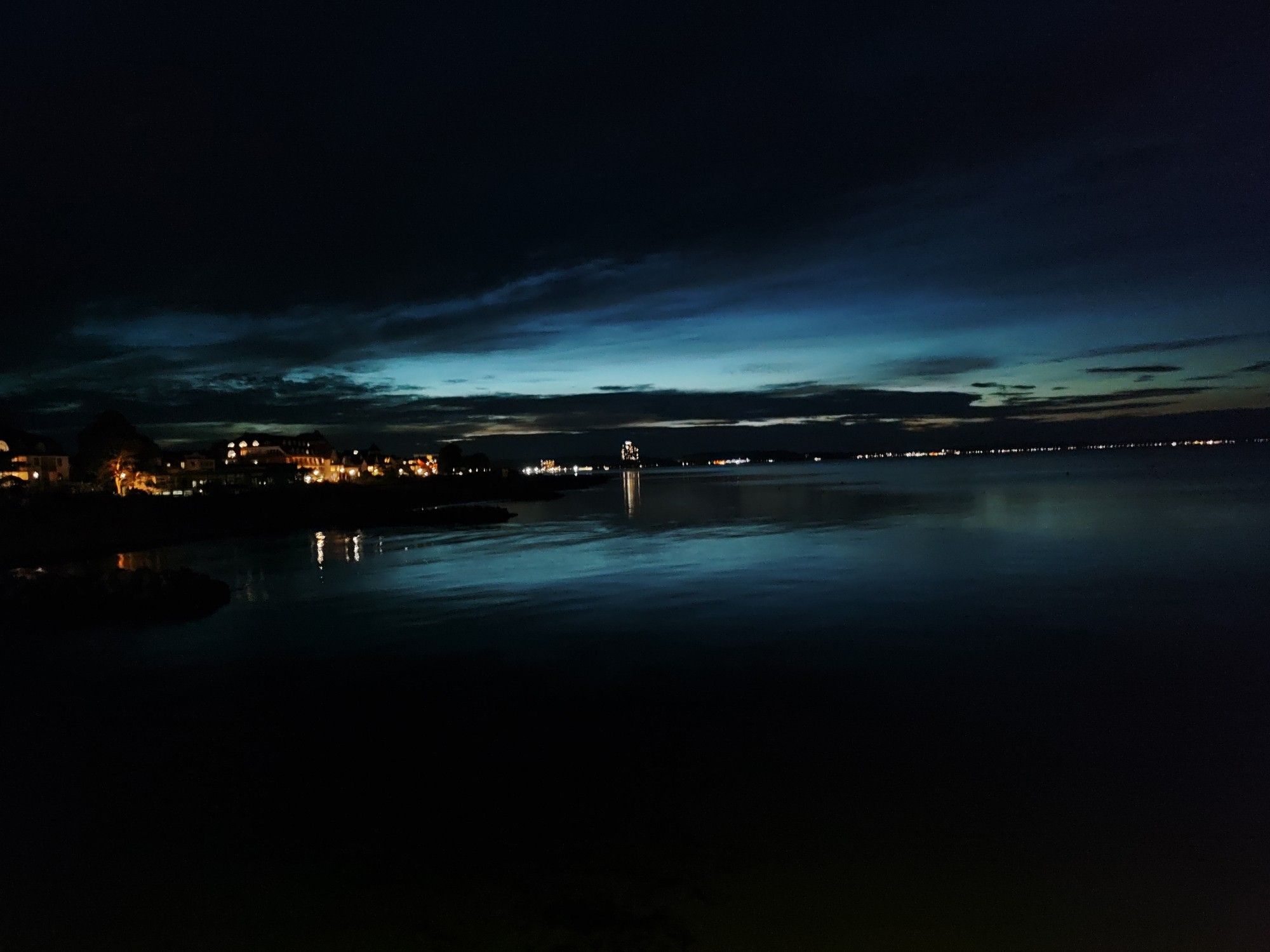 Abends an der Ostsee, der blauschwarze Himmel spiegelt sich im Meer. Die Lichter am Ufer leuchten.