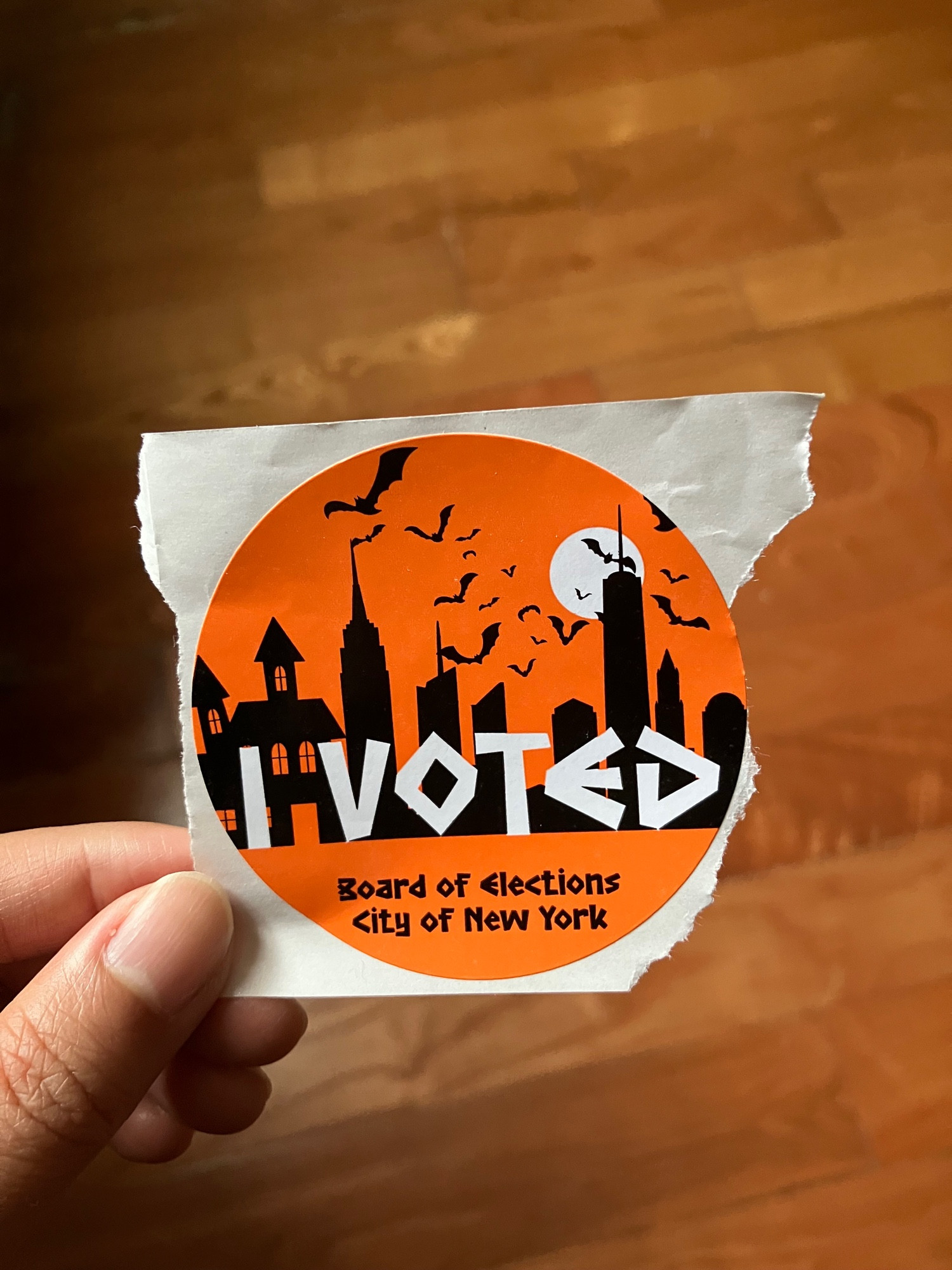 An “I VOTED” sticker that is Halloween themed, with an orange background, a black NYC skyline with spooky vibes, bats flying around the skyline, and a full moon