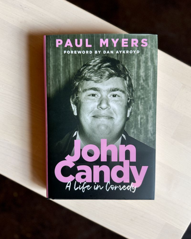 A hardcover copy of 'John Candy: A Life in Comedy' featuring a smiling, black and white image of John Candy with bold, pink text, sits on a light wooden surface. 