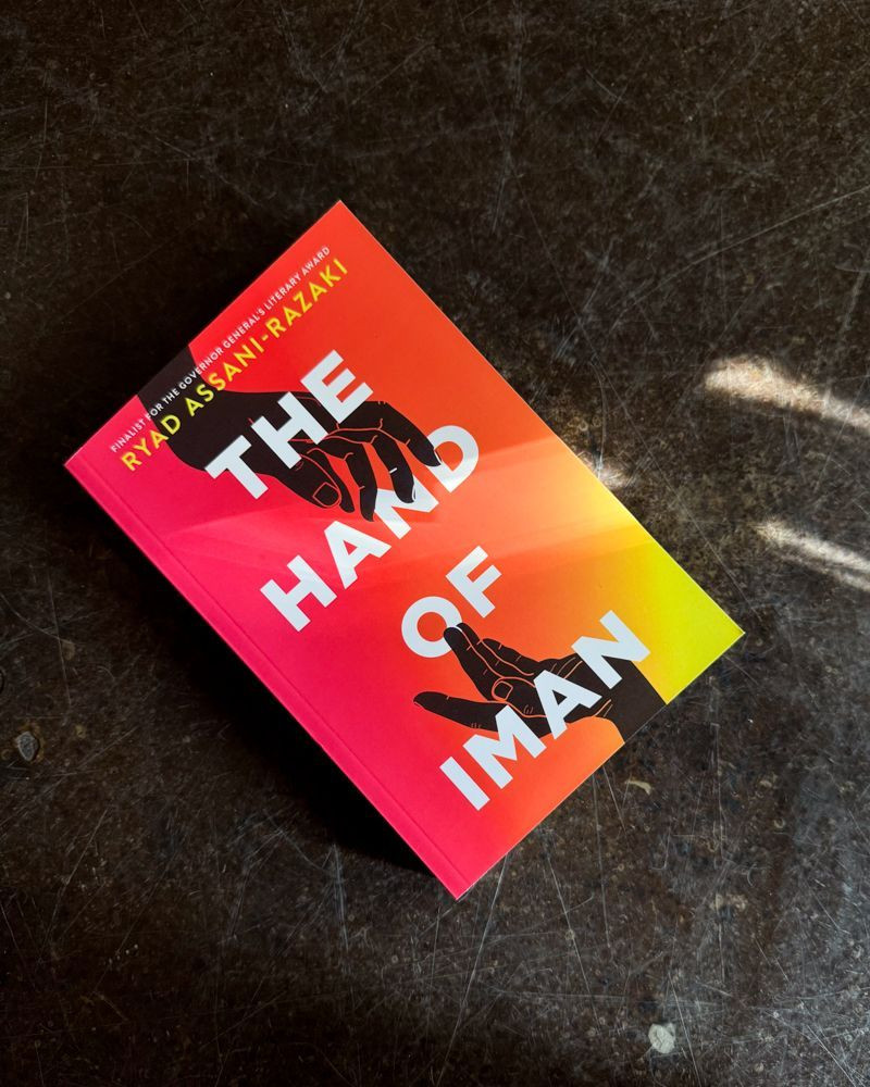 A copy of THE HAND OF IMAN sits on a concrete floor with shadows cast across it. The cover features a gradient red to yellow, with two hands reaching towards each other.