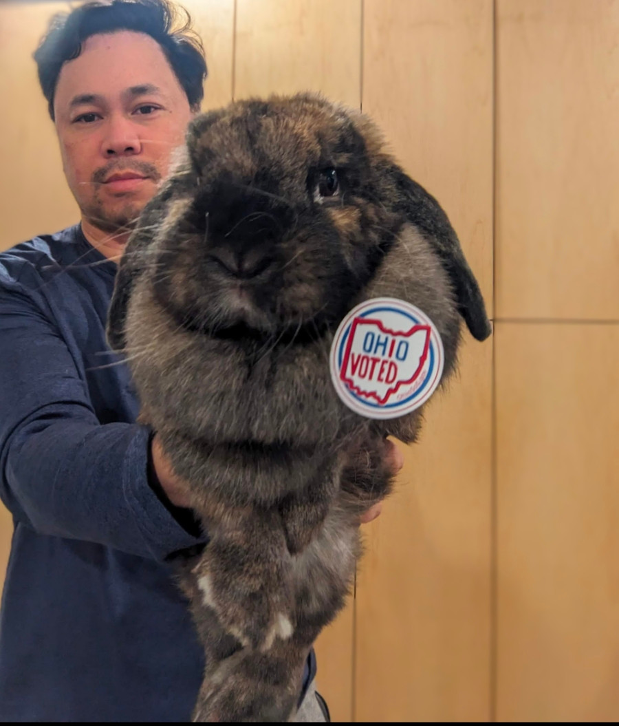 Me a middle aged Asian male in  blue shirt holding up a brown lop eared bunny with an "Ohio voted" sticker on his dewlap. 