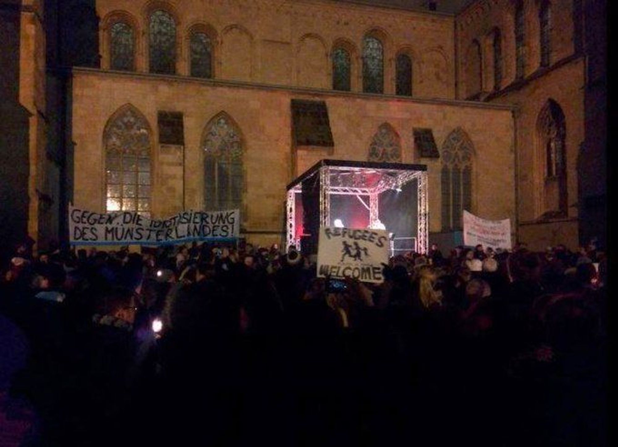 Eine große Menge von Demonstrant*innen versammelt sich in der Dunkelheit vor einer Bühne an der Lambertikirche in Münster. Es sind mehrere Transparente dabei, u.a. eins mit dem Slogan "Refugees Welcome".