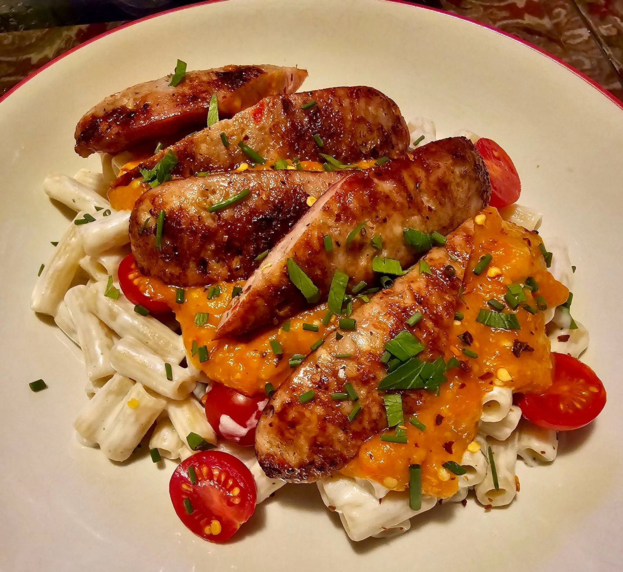 A plate of pasta topped with a creamy orange pepper sauce, halved cherry tomatoes and slices of chicken Italian sausage.