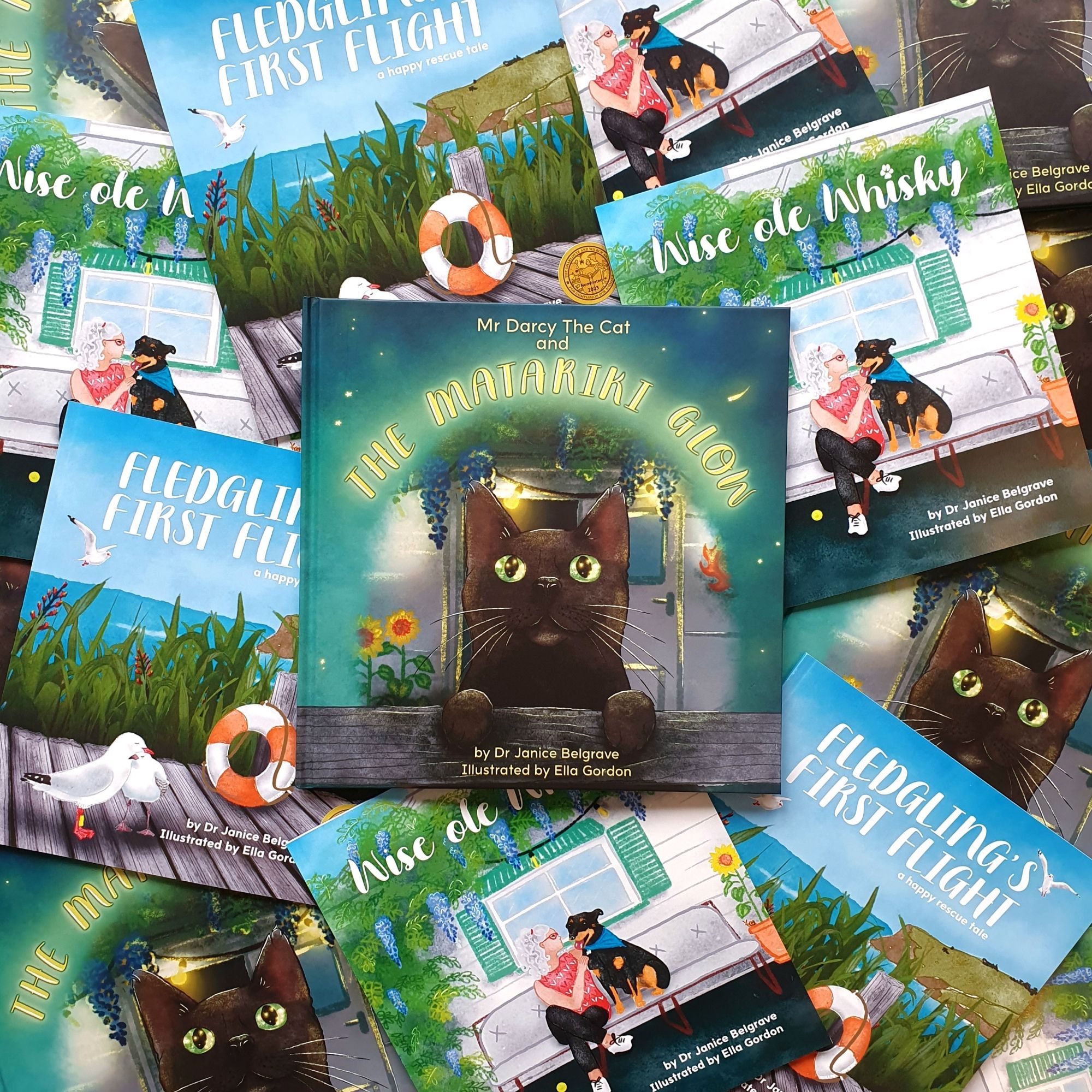 A range of children's picture books are spread across q flat surface. The book in the middle is titled Mr Darcy The Cat and The Matariki Glow by Dr Janice Belgrave Illustrated by Ella Gordon. The front cover is an illustration of a black cat looking up to the stars. Other kid lit books lay around it with covers including an array of birds, dogs and people.