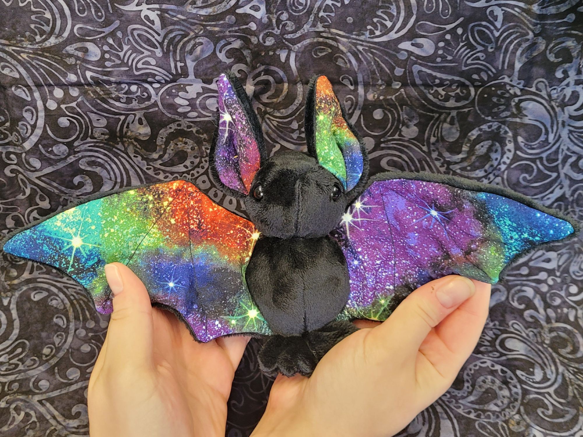 a picture of a bat plushie held up in front of a black background. The bat has a black minky body, with wings outspread wide. The cotton fabric on his wings and inside his ears is a mottled rainbow colored galaxy and white stars.