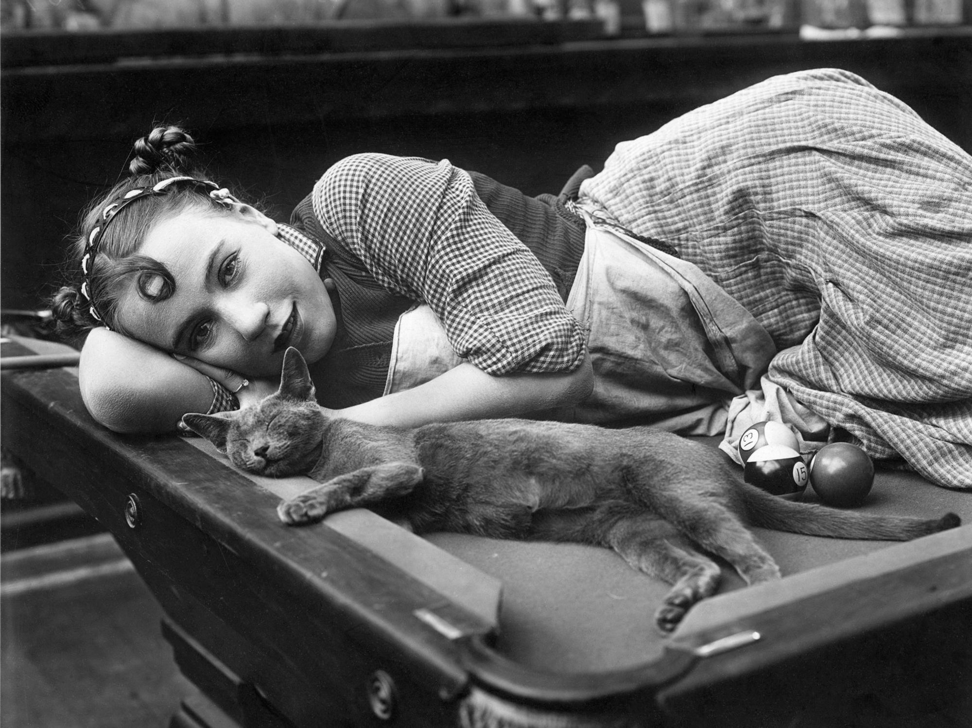 Photo en noir et blanc d'une jolie jeune femme, souriante, portant une robe à carreaux, couchée sur une table de billard derrière un jeune chat sans doute gris, ravissant, les yeux fermés.