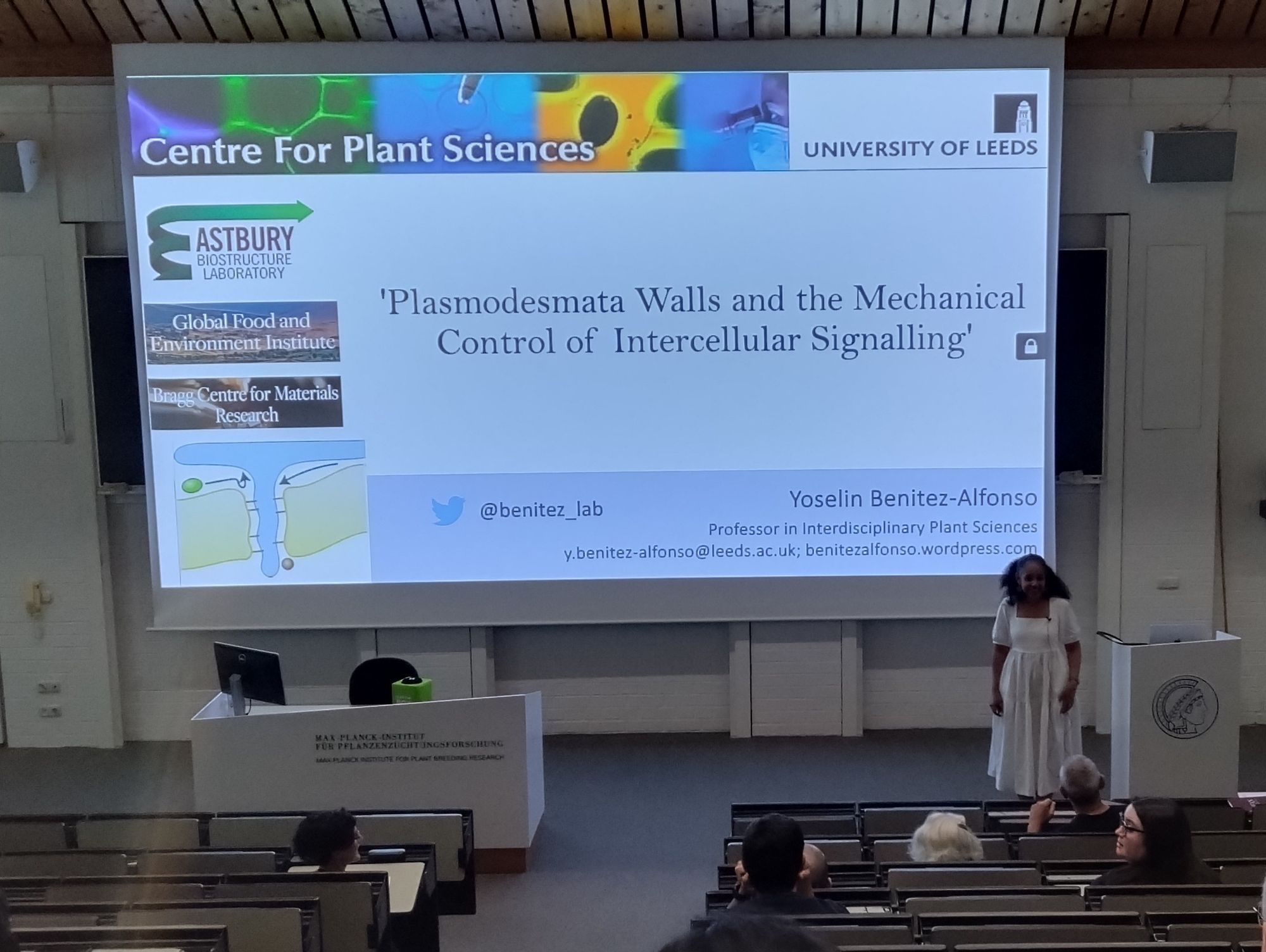 A picture of Yoselin Benitez-Alfonso on stage in the lecture hall of the Max Planck Institute for Plant Breeding Research in Cologne. Her slide shows the title "Plasmodesmata Walls and the Mechanical Control of Intercellular Signalling".