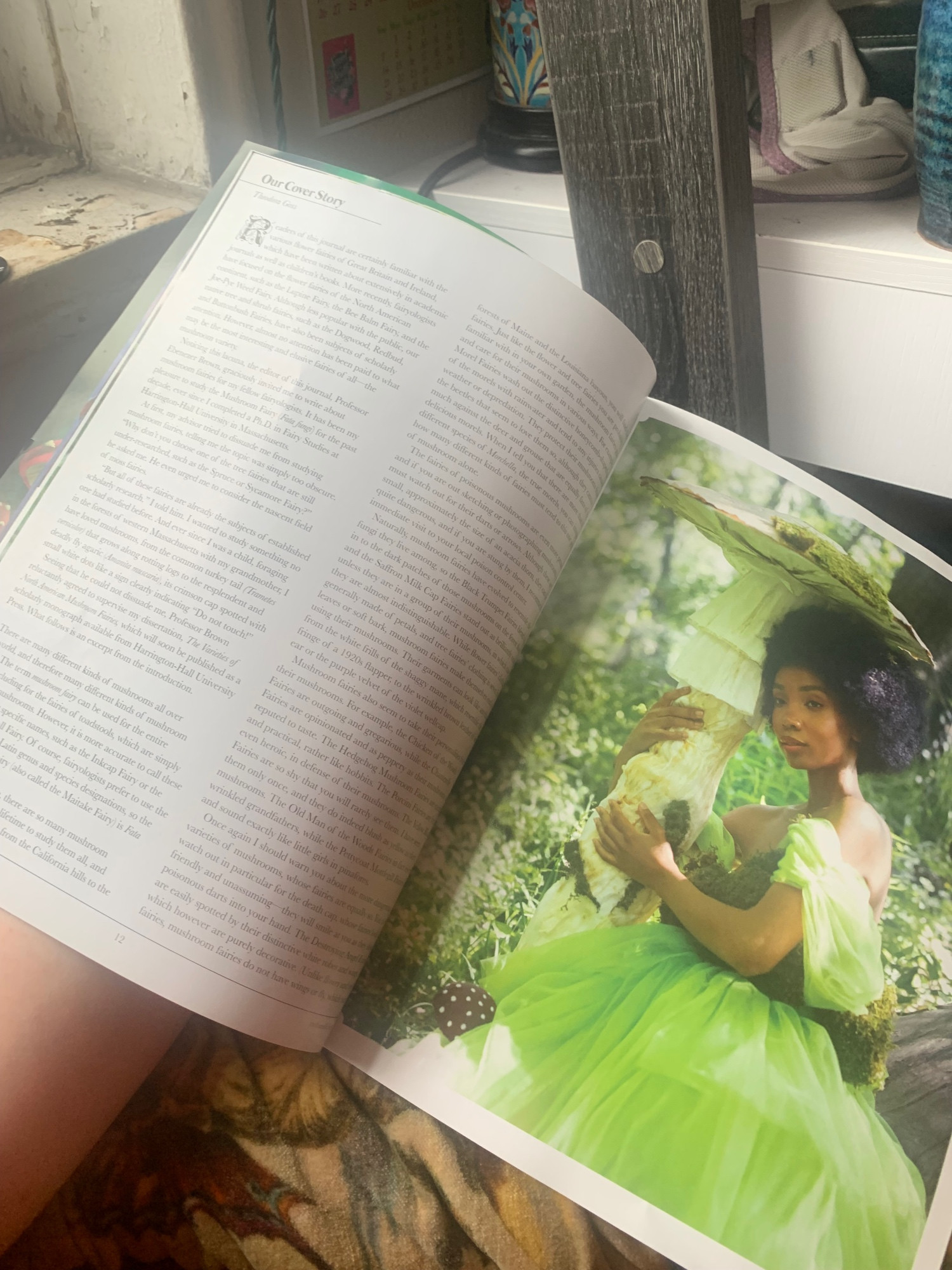 An image of Enchanted Living Magazine Issue 70. A beautiful black woman in a bright green tulle gown embraces a forest tree trunk, surrounded by mist and leaves and ferns and mushrooms. 