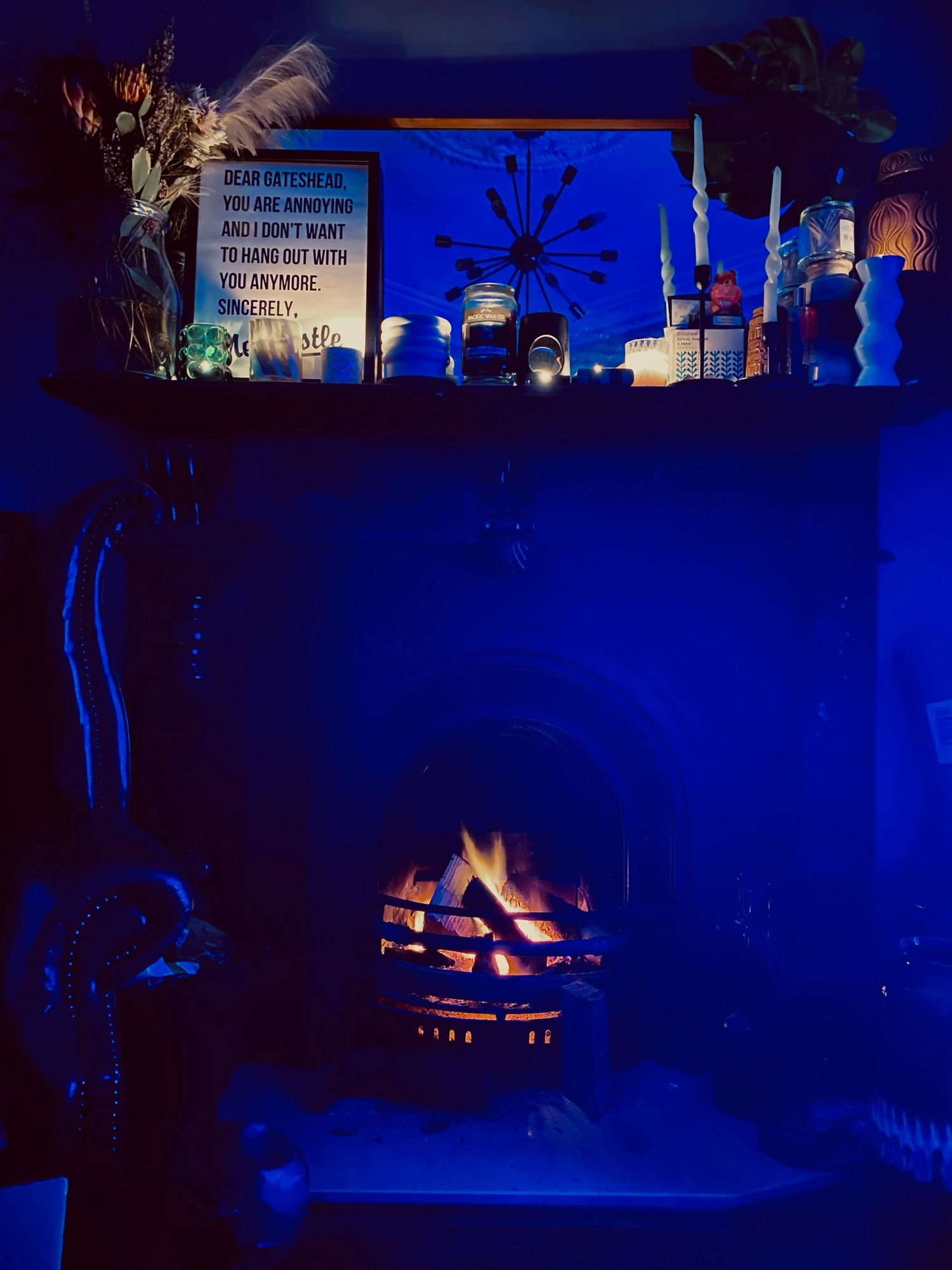 Fireplace with glowing fire- Mantle has lots of different size candles on and a poster saying ‘dear Gateshead you are annoying and I don’t want to hang out with you anymore, sincerely Newcastle’.