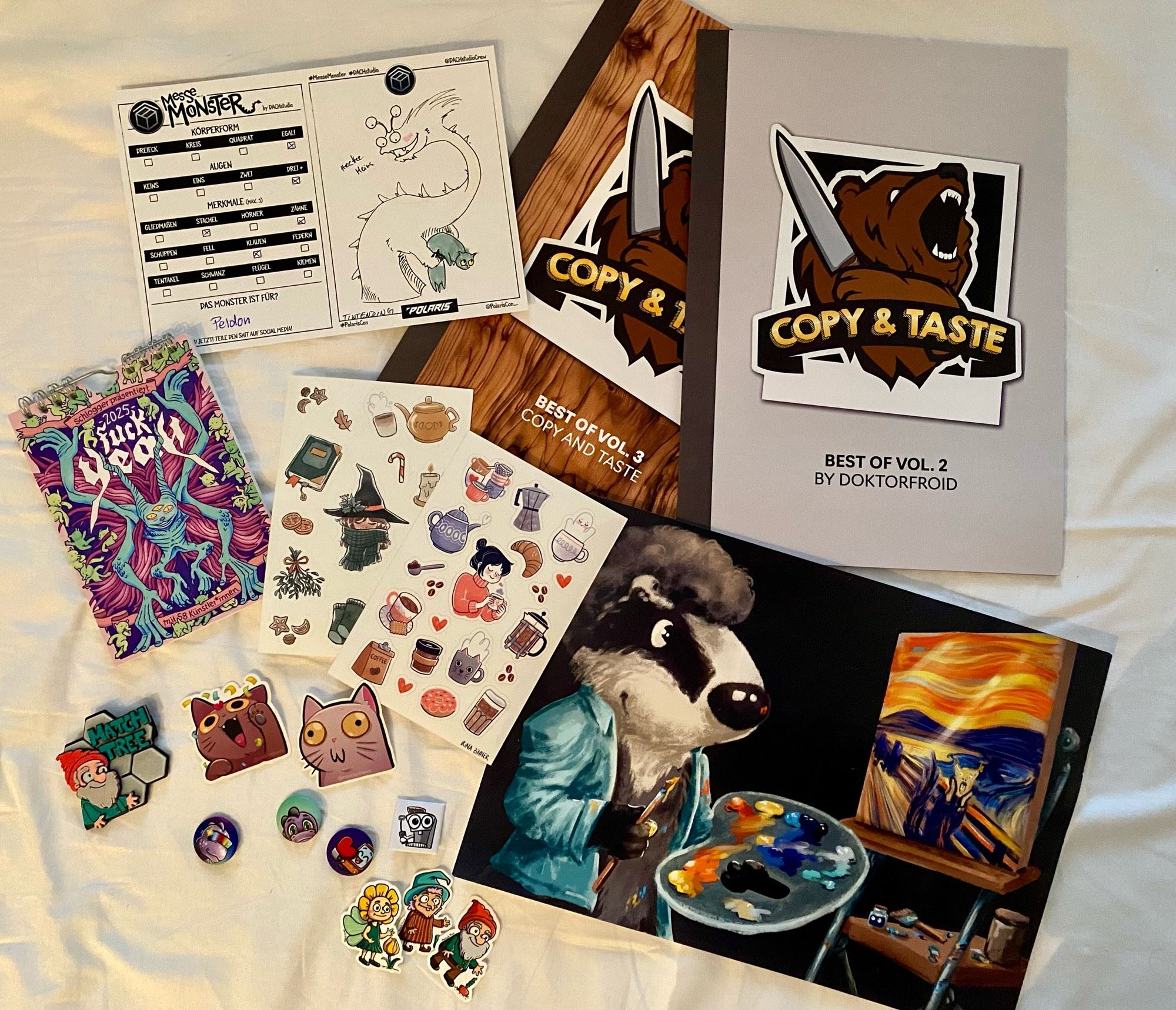 Verschiedenste Sticker und Buttons. Ein Kunst Kalender, ein Artwork, 2 Kochhefte und ein Messemonster. 