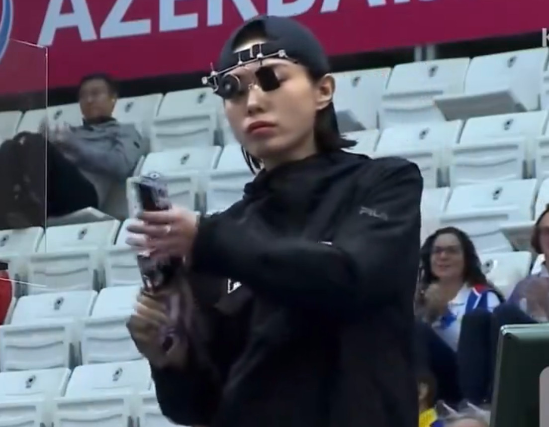 kim yeji at the 2024 olympics reloading a gun with the korean flag on it, wearing a backwards black snapback and steampunk looking little round sunglasses that look like they help her aim