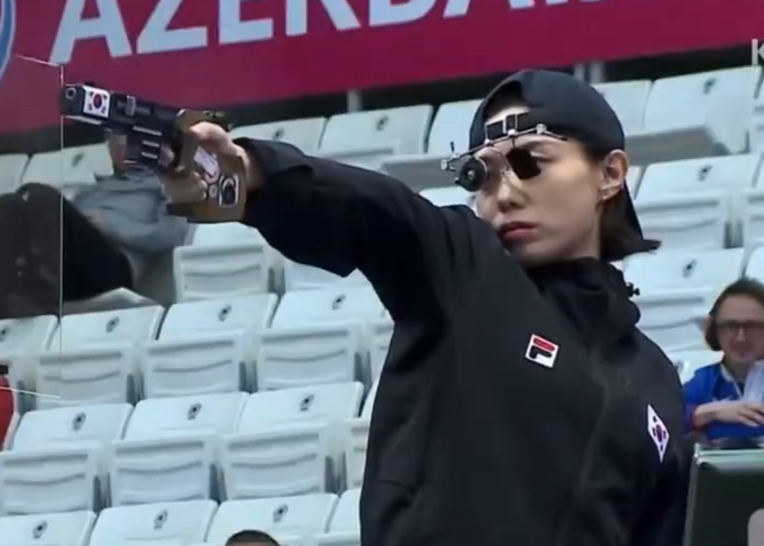 kim yeji at the 2024 olympics aiming a gun with the korean flag on it, wearing a backwards black snapback and steampunk looking little round sunglasses that look like they help her aim