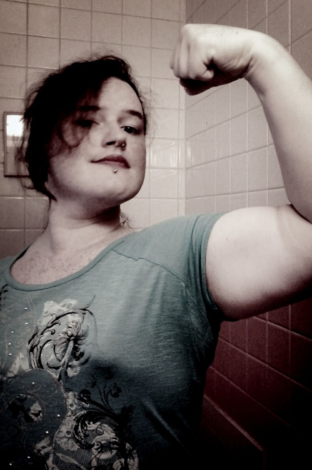 the other picture from the muscle flexing set. a woman with hair in a ponytail you can't really see but some of which is spilling over one side of her face is smiling lifting her head while looking down towards the camera. she is flexing her bicep which is being cut into a bit by the overly tight teal shirt