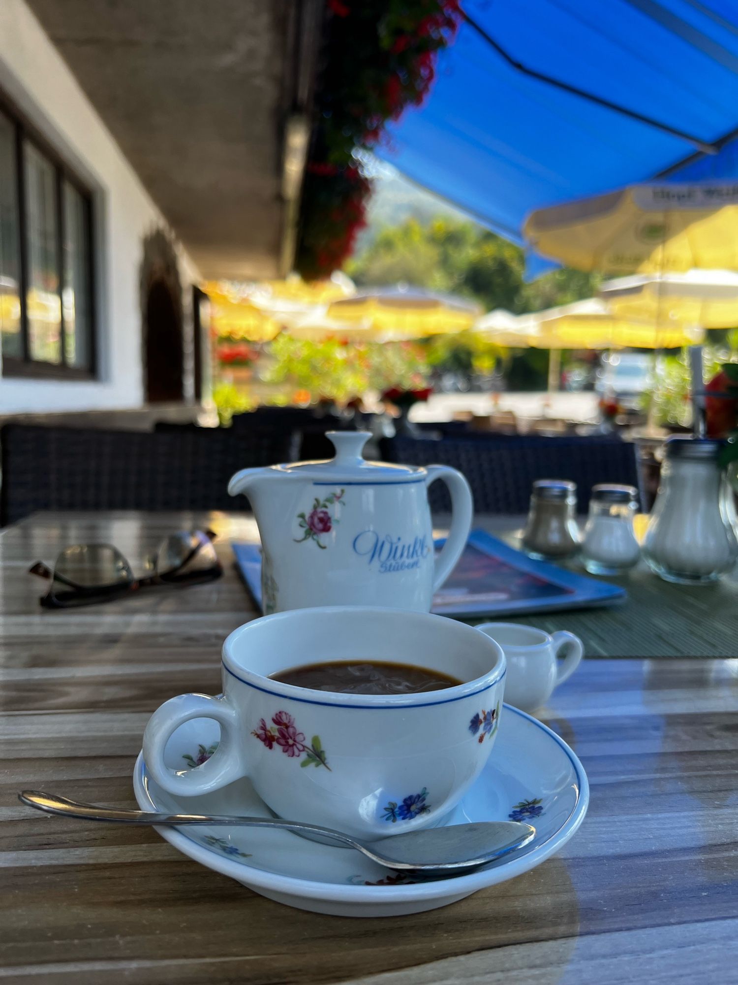 Ein Kännchen und eine Tasse Kaffee in weißem, mit kleinen Blüten verzierten Porzellan steht auf einem Tisch in der Außengastro