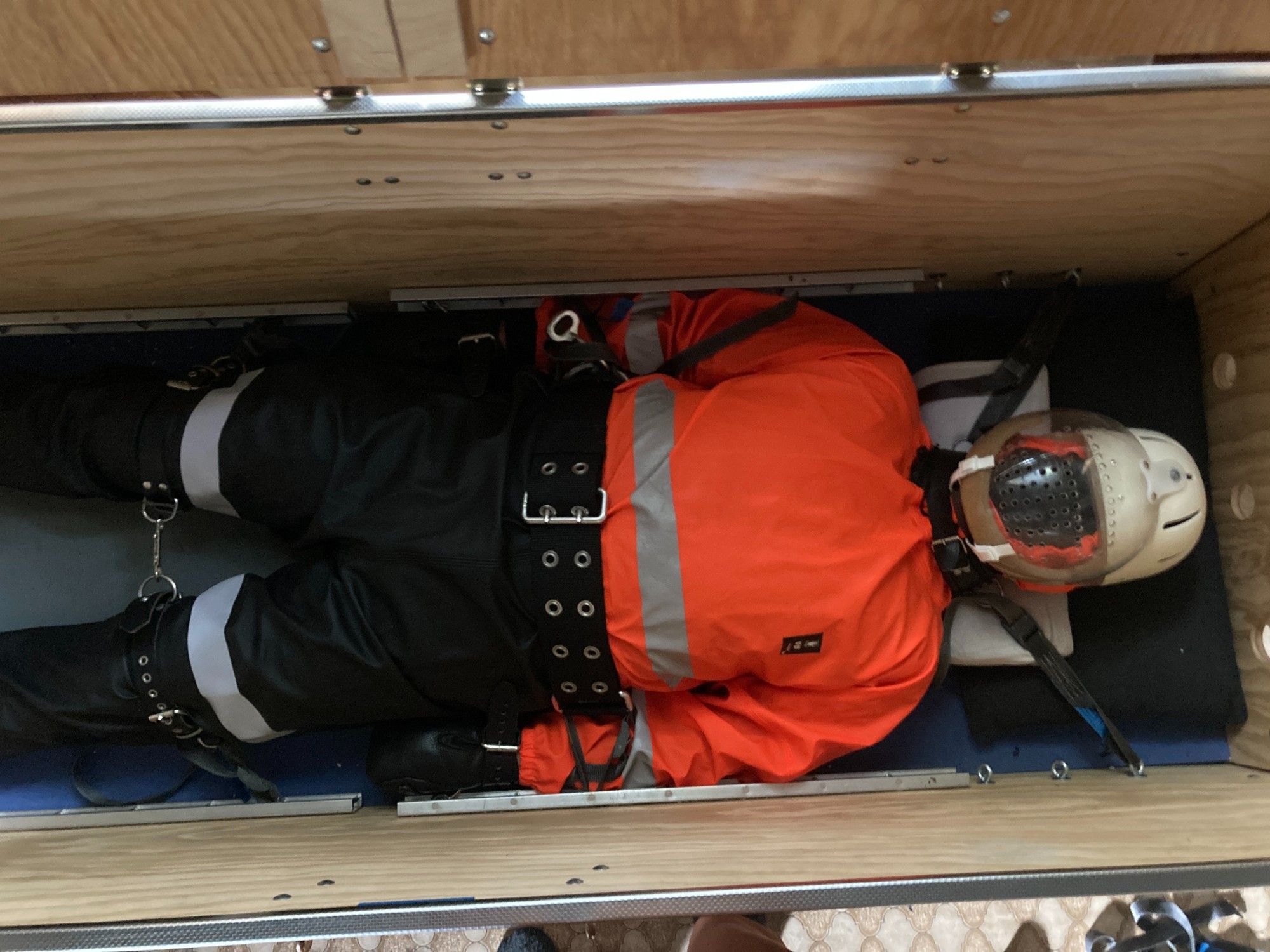 Me in a hi-viz wading suit wearing a Danmar helmet, Sinvention mitts, and a firefighters belt, limbs and neck strapped inside a wooden box.