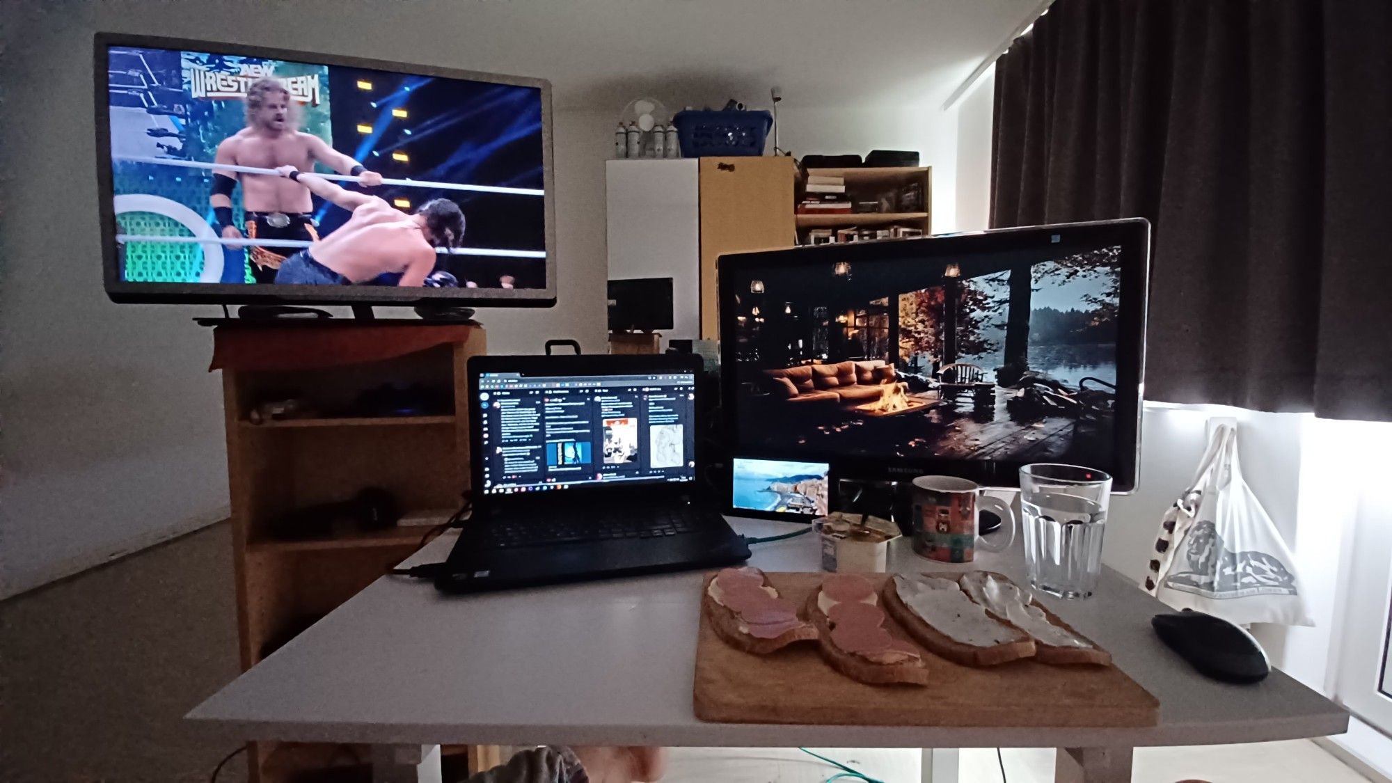 Bild meines Schreibtisches. Darauf ein Brett mit belegten Broten,Kaffeetasse,ein Joghurt,Desktop und Laptop. Auf dem Fenrseher links läuft der Wrestling PPV AEW Wrestledream.