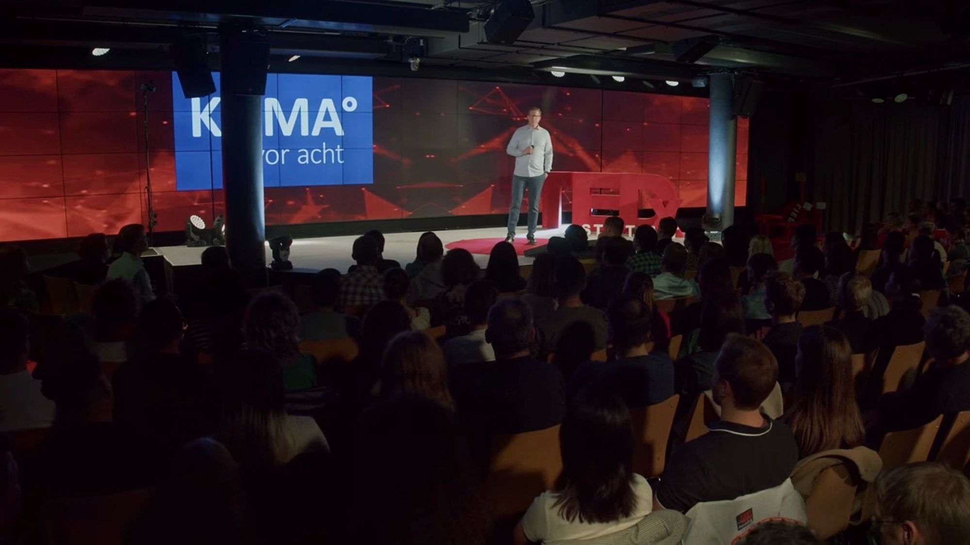 Norman Schumann von KLIMA° vor acht auf der TedX Bühne in Stuttgart. Auf der Videowand ist das Logo von Klima vor acht zu sehen. Im Vordergrund sitzen viele Zuschauer:innen im abgedunkelten Saal.