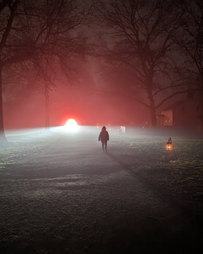 a nighttime photo with silhouette of a child walking down a dirt road in the fog, with trees on either side of the road, and a bright light off in the distance (creating the silhouette and shadow stretching out in front of the child). there is a creepy looking house on the right, lit only by candles, and a few candle-lit lanterns off in the distance.