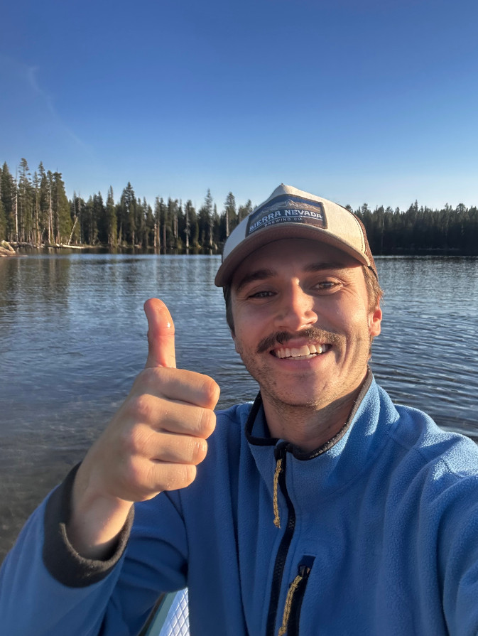 Photo of a guy in a kayak or canoe smiling and giving a thumbs up. He has a mustache, and a blue zip fleece, and is wearing a Sierra Nevada hat.